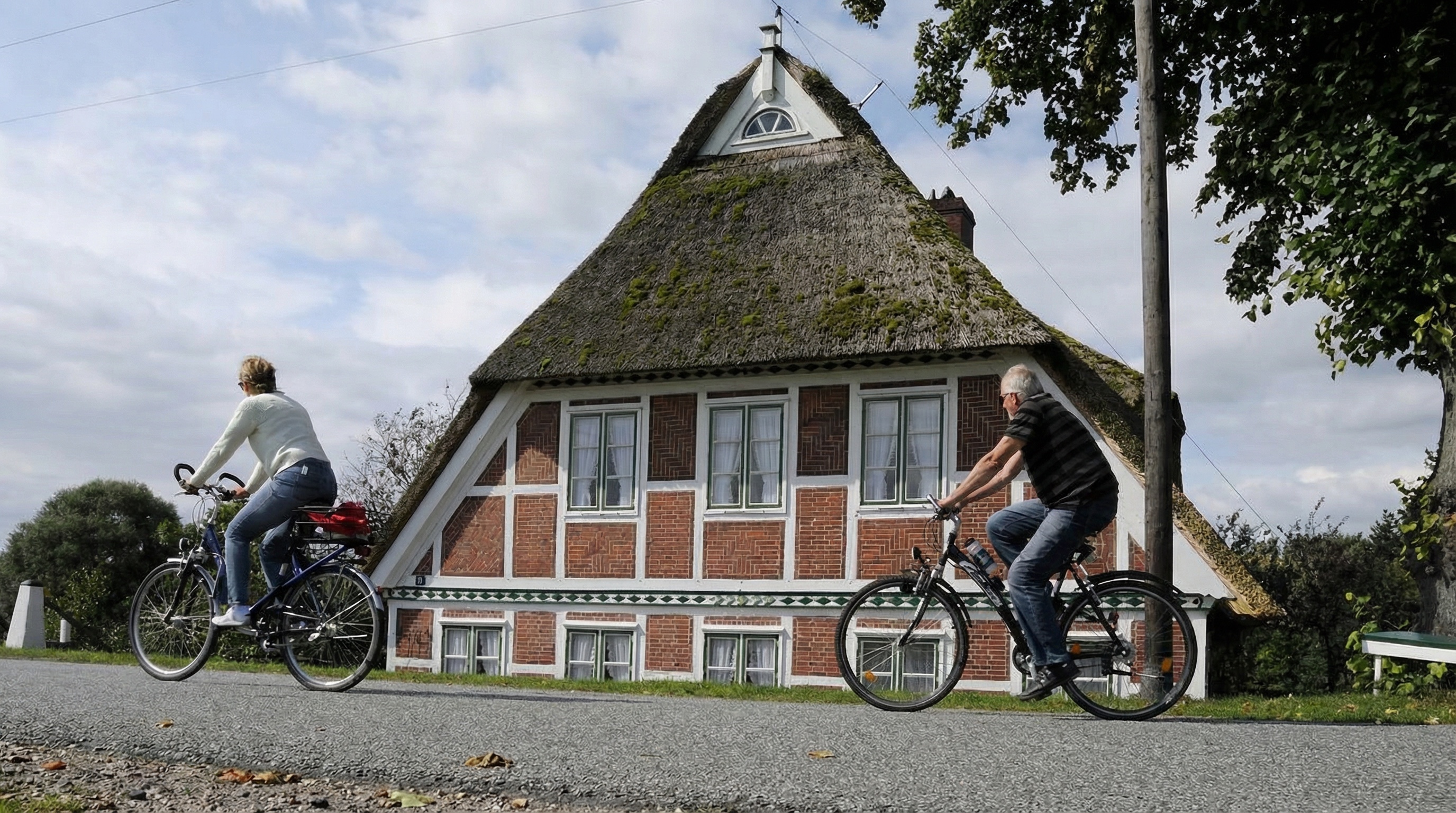 Zwei Radfahrer fahren an einem Fachwerkhaus mit Reetdach und mehreren Fenstern vorbei, im Hintergrund Bäume und bewölkter Himmel in Altengamme, Hamburg.