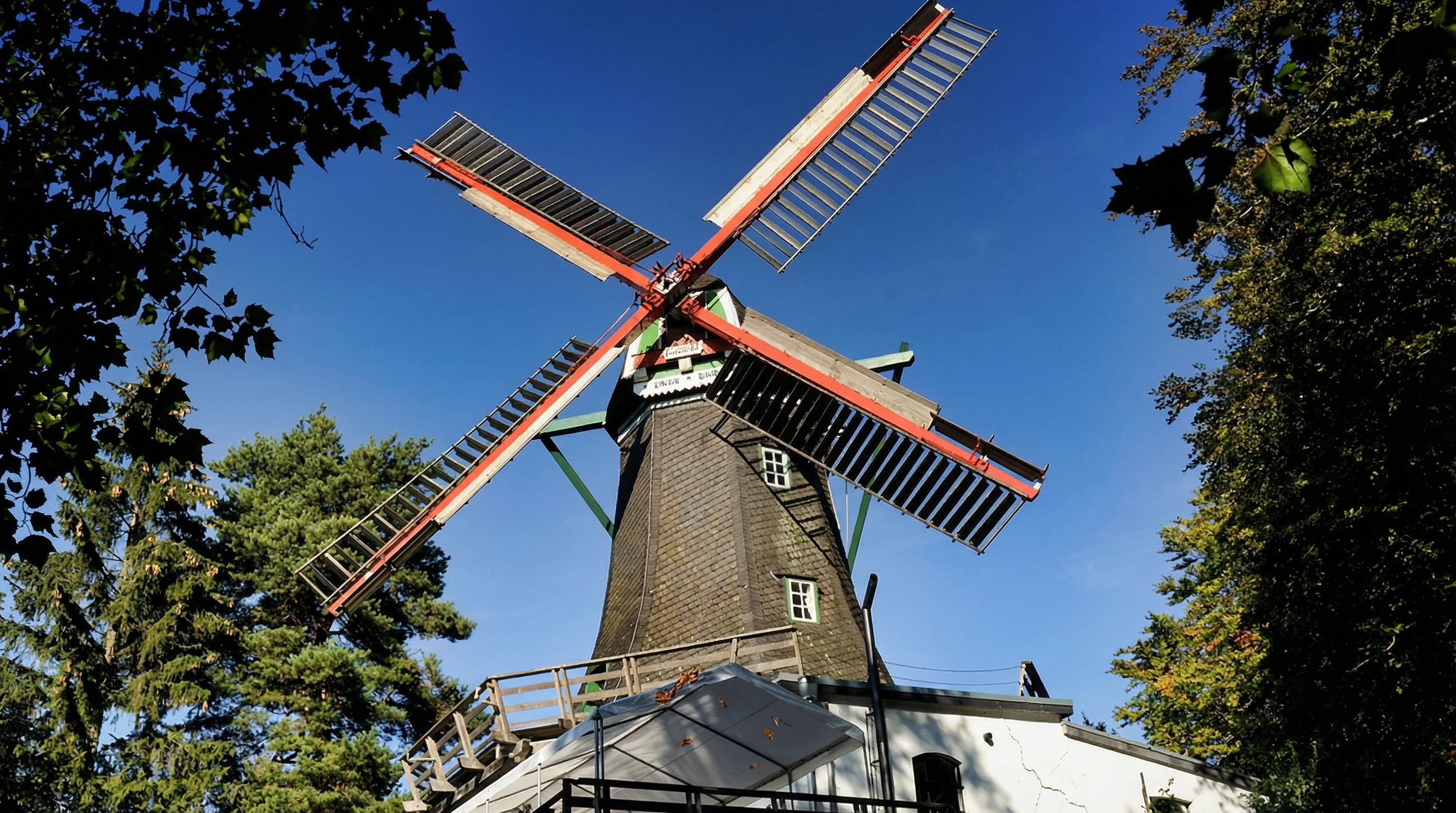 Windmühle mit rot-weißen Flügeln, braunem Turm und weißem Sockel, umgeben von grünen Bäumen unter blauem Himmel in Bergedorf, Hamburg.