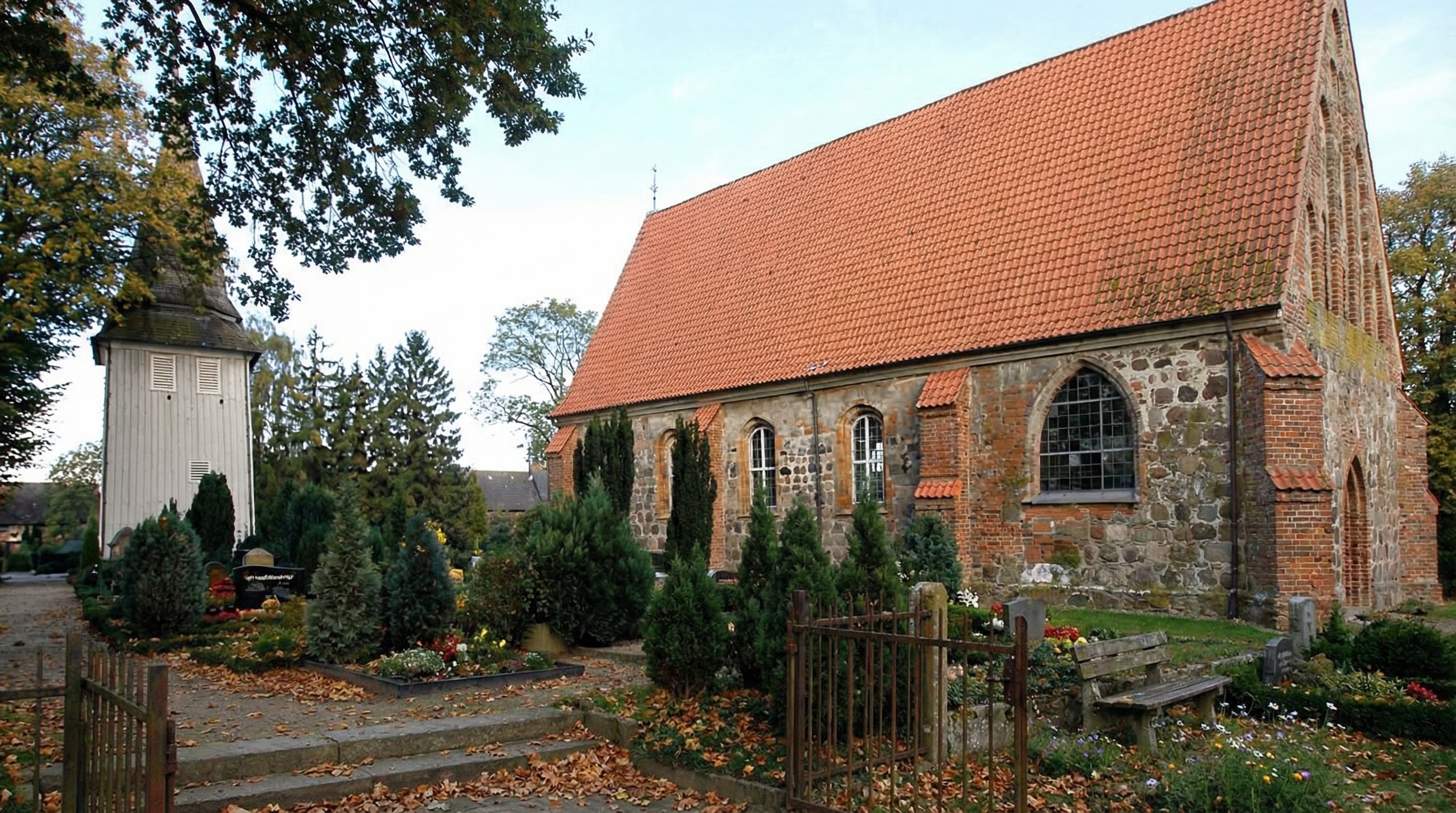 Kirche aus Feldsteinen mit rotem Ziegeldach, daneben hölzerner Glockenturm, umgeben von Bäumen und gepflegtem Friedhof mit Gräbern in Neuengamme, Hamburg.