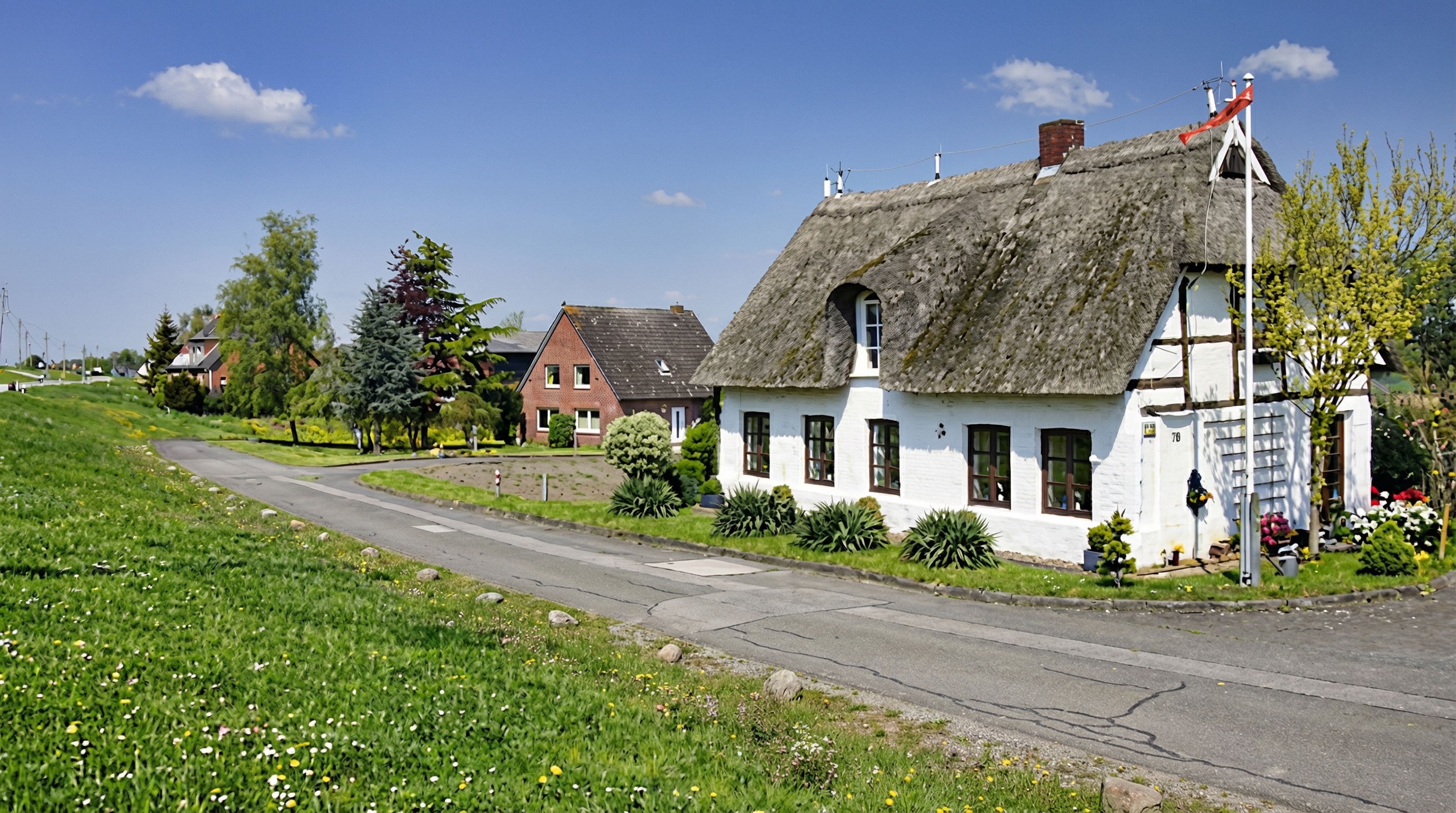 Idyllische Dorfszene mit einem weissen Reetdachhaus im Vordergrund, umgeben von gepflegtem Garten und Pflanzen. Im Hintergrund stehen weitere Backsteinhäuser entlang einer schmalen, gewundenen Strasse. Rechts neben dem Haus weht eine Fahne an einem Mast. Die Landschaft ist grün und blühend unter einem klaren, blauen Himmel in Spadenland, Hamburg.