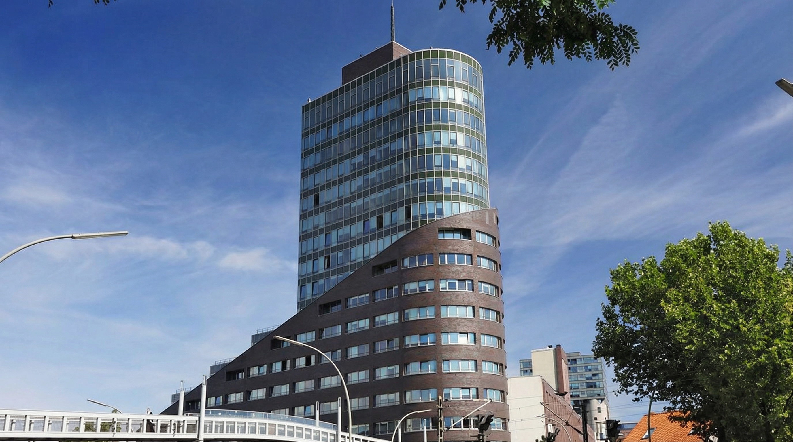 Modernes Hochhaus mit markanter Architektur, bestehend aus einer unteren Etage mit brauner Klinkerfassade und oberen Stockwerken mit Glasfassade. Das Gebäude hat eine geschwungene Form und erhebt sich über eine Stadtlandschaft mit Strassenlaternen, Bäumen und weiteren Gebäuden im Hintergrund. Der Himmel ist klar mit leichten Wolkenstreifen in Harburg, Hamburg.