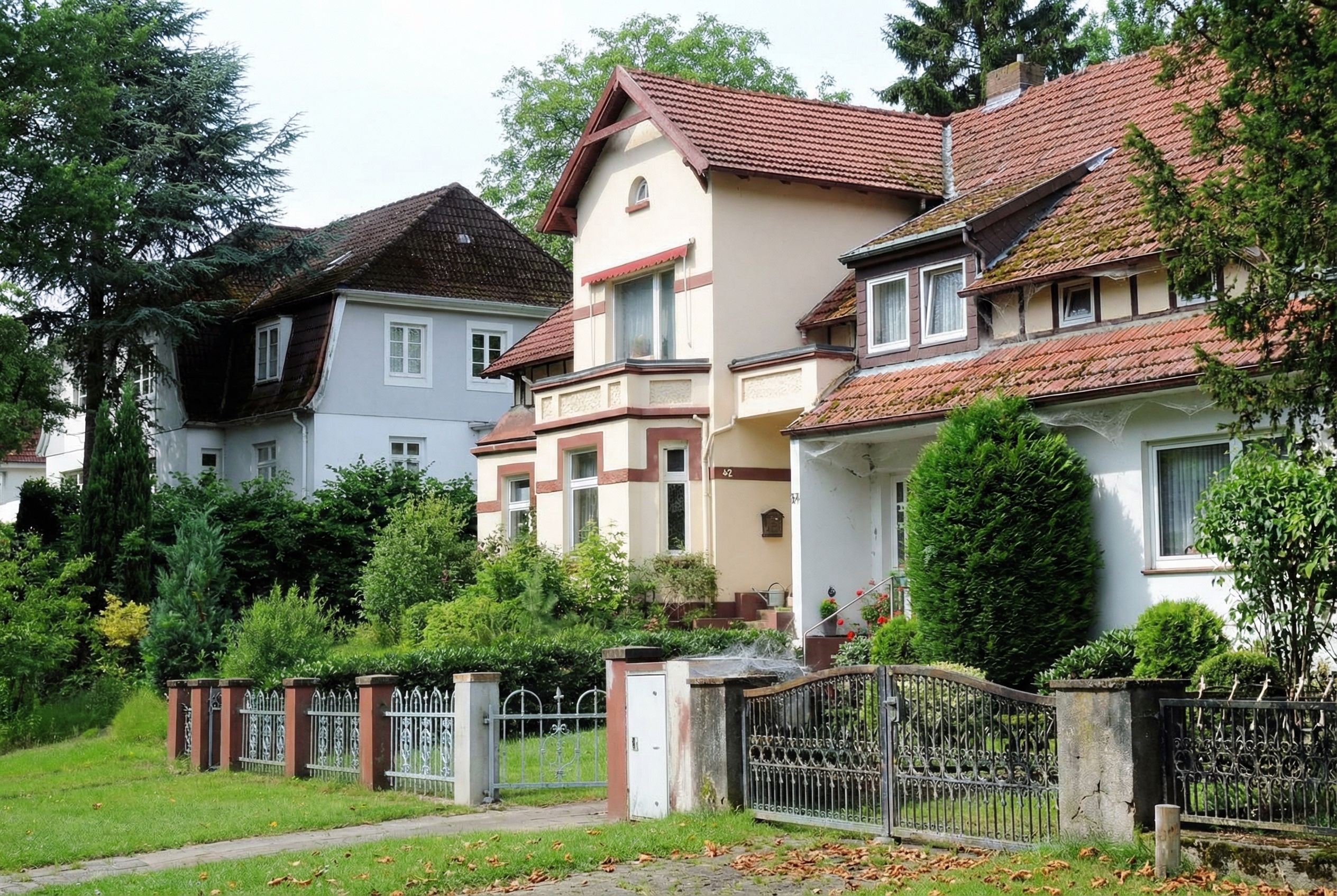 Mehrere ältere Einfamilienhäuser mit Ziegeldächern in einer ruhigen Wohngegend, teils mit Erkern und Fachwerkdetails. Die Häuser sind von gepflegten Gärten mit Büschen, Hecken und kleinen Bäumen umgeben. Im Vordergrund verläuft ein Gehweg mit einem schmiedeeisernen Zaun und geöffnetem Einfahrtstor. Herbstlaub liegt verstreut auf dem Rasen und Gehweg in Eißendorf, Hamburg.