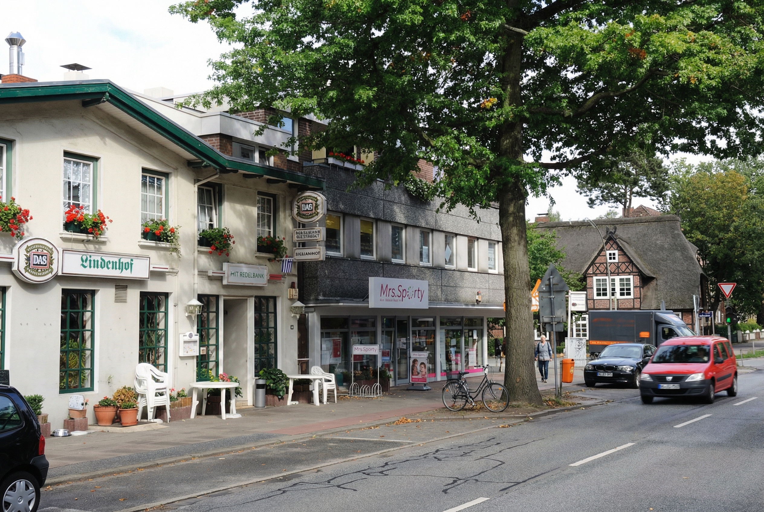 Belebte Strassenszene mit mehreren Gebäuden, darunter ein traditionelles Gasthaus namens "Lindenhof" mit Blumenkästen an den Fenstern, ein modernes Fitnessstudio "Mrs. Sporty" im Nachbargebäude sowie ein Fachwerkhaus mit Reetdach im Hintergrund. Autos und ein Lieferwagen fahren auf der Strasse, ein Fahrrad steht am Gehweg, und Fussgänger sind unterwegs. Ein grosser Baum spendet Schatten in Heimfeld, Hamburg.