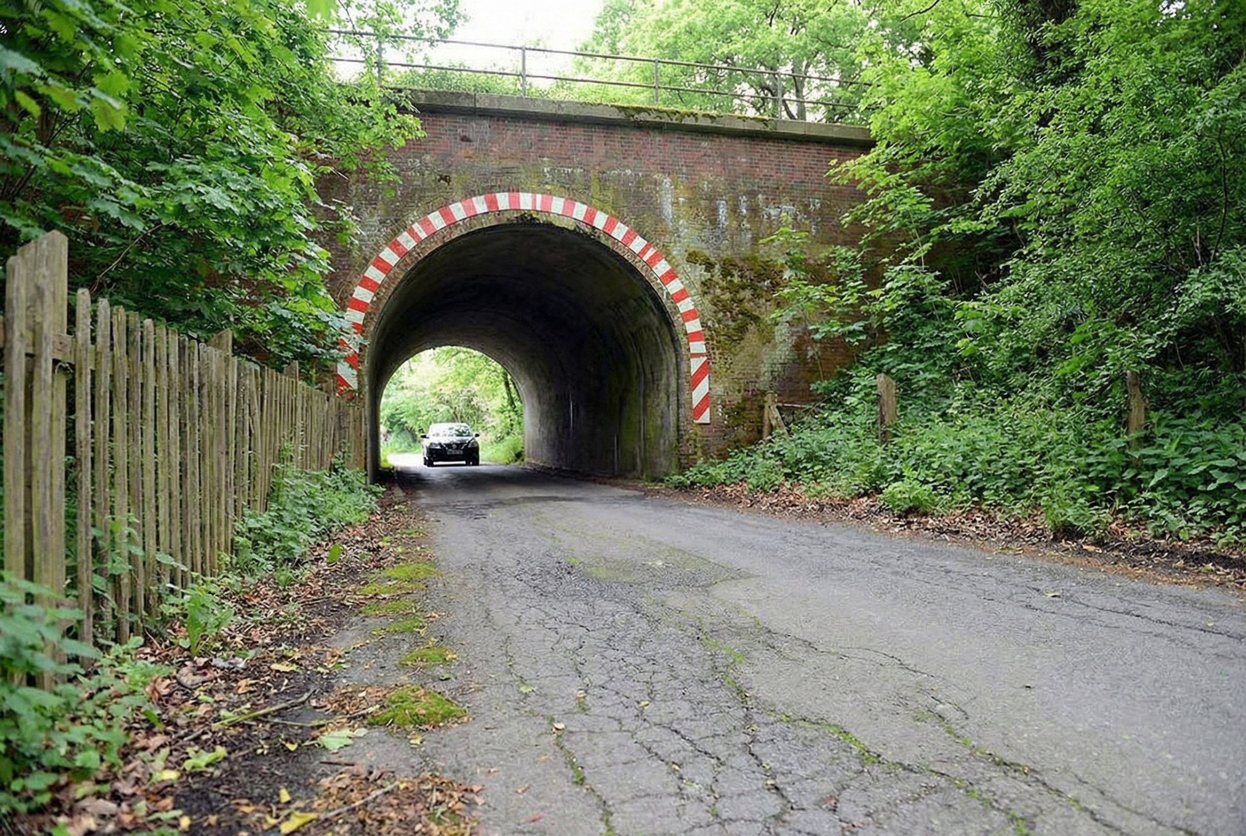 Schmale, von Bäumen gesäumte Landstrasse mit Rissen im Asphalt, die durch einen alten gemauerten Tunnel mit rot-weiss markiertem Rundbogen führt. Ein Auto fährt aus dem Tunnel, links verläuft ein Holzzaun, rechts dichter Bewuchs. Die Szene wirkt ruhig und ländlich in Rönneburg, Hamburg.