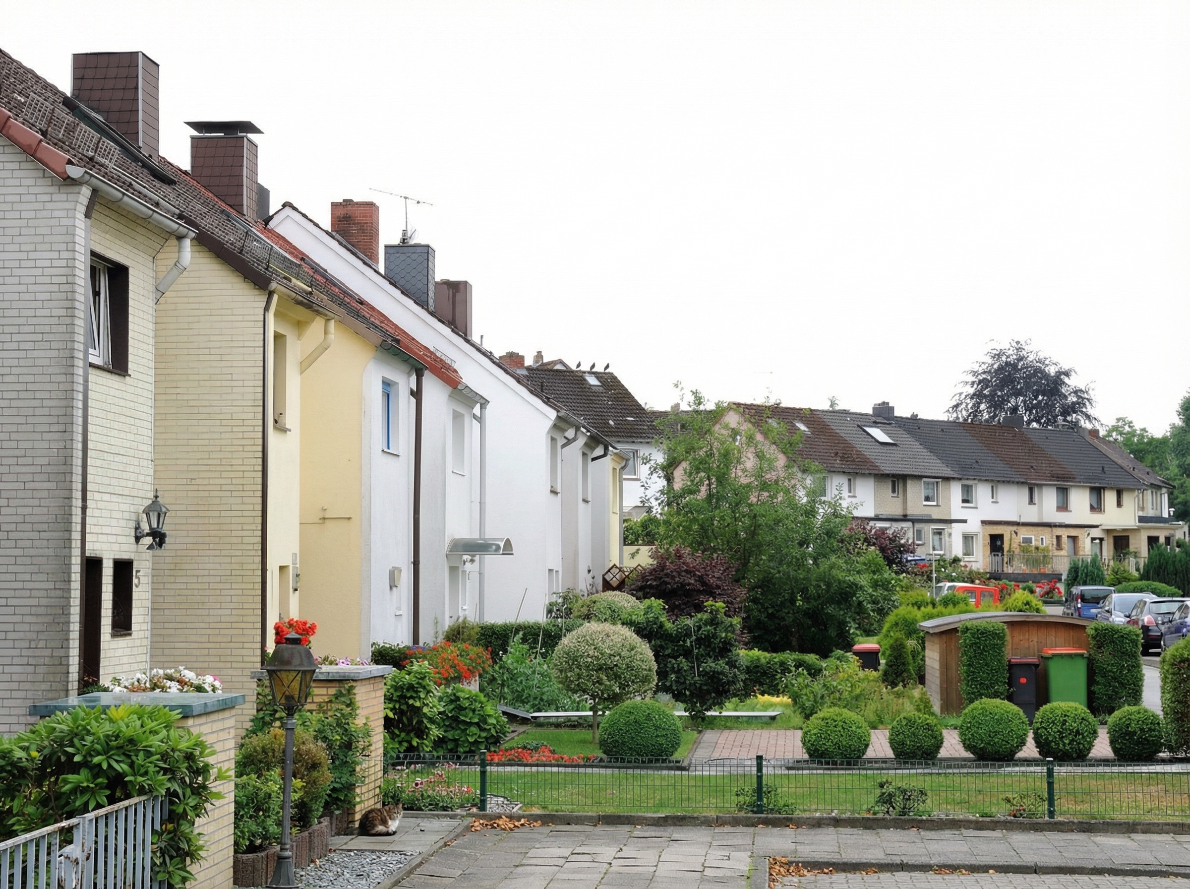 Reihenhaus-Siedlung mit gepflegten Vorgärten, kleinen Rasenflächen und Ziersträuchern. Die Häuser sind in hellen Farben gestrichen und haben Satteldächer mit Dachfenstern und Schornsteinen. Im Vordergrund befinden sich ein kleiner Weg, eine Laterne, eine sitzende Katze sowie ein Gartenbereich mit Müll in Langenbek, Hamburg.