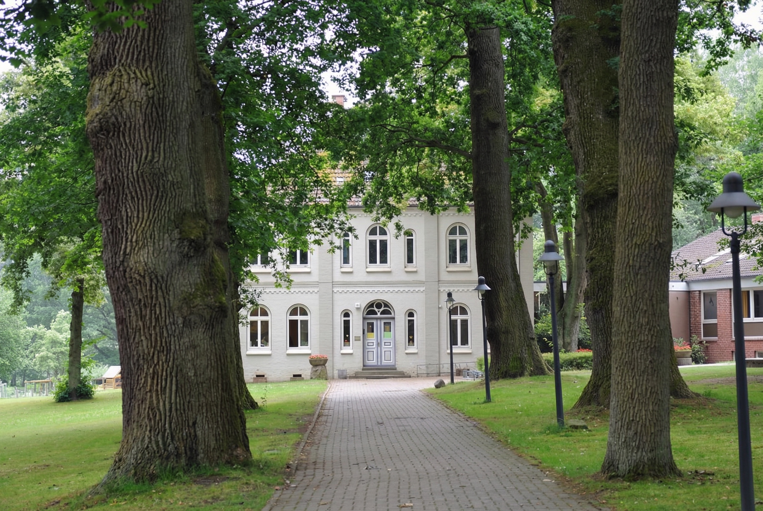 Gepflasterter Weg, gesäumt von alten, hohen Bäumen, führt zu einem zweistöckigen, hell gestrichenen Herrenhaus mit symmetrischer Fassade und grossen Rundbogenfenstern. Auf dem Rasen stehen mehrere Laternen, rechts im Hintergrund ist ein modernes Backsteingebäude zu sehen. Die Umgebung wirkt gepflegt und parkähnlich in Sinstorf, Hamburg.