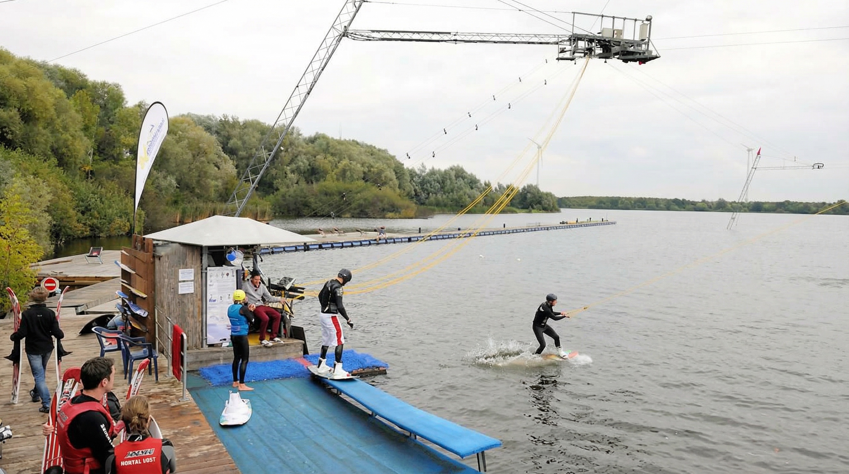 Wasserskianlage an einem See mit Seilzugsystem, das mehrere Zugseile über das Wasser spannt. Im Vordergrund startet eine Person mit Wakeboard von einer blauen Rampe, eine weitere steht bereit, während andere Teilnehmende mit Wasserskiern am Steg warten. Ein Mitarbeiter bedient die Anlage in einer kleinen Holzhütte. Links ist ein Holzsteg mit Stühlen und Wasserskiausrüstung zu sehen, im Hintergrund dichter Baumbewuchs, ein langer Schwimmsteg im See und mehrere Windräder am Horizont in Neuland, Hamburg.