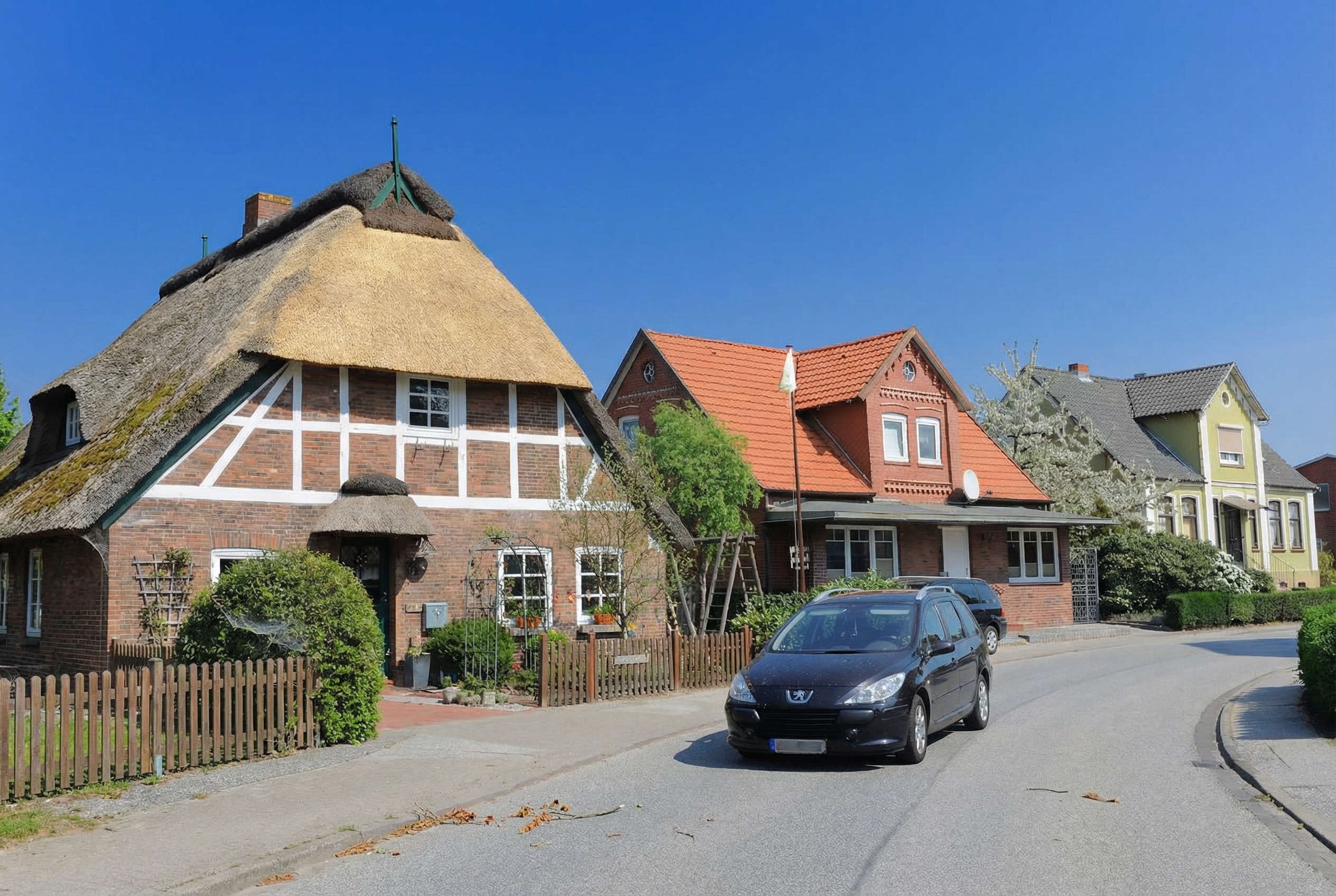 Strassenszene mit mehreren Einfamilienhäusern in unterschiedlichen Baustilen, darunter ein Fachwerkhaus mit neuem Reetdach, ein Backsteinhaus mit rotem Ziegeldach und ein hellgrünes Haus mit klassischer Fassade. Vor den Häusern stehen gepflegte Vorgärten, auf der Strasse ist ein geparktes Auto zu sehen. Der Himmel ist klar und blau in Neuenfelde, Hamburg.