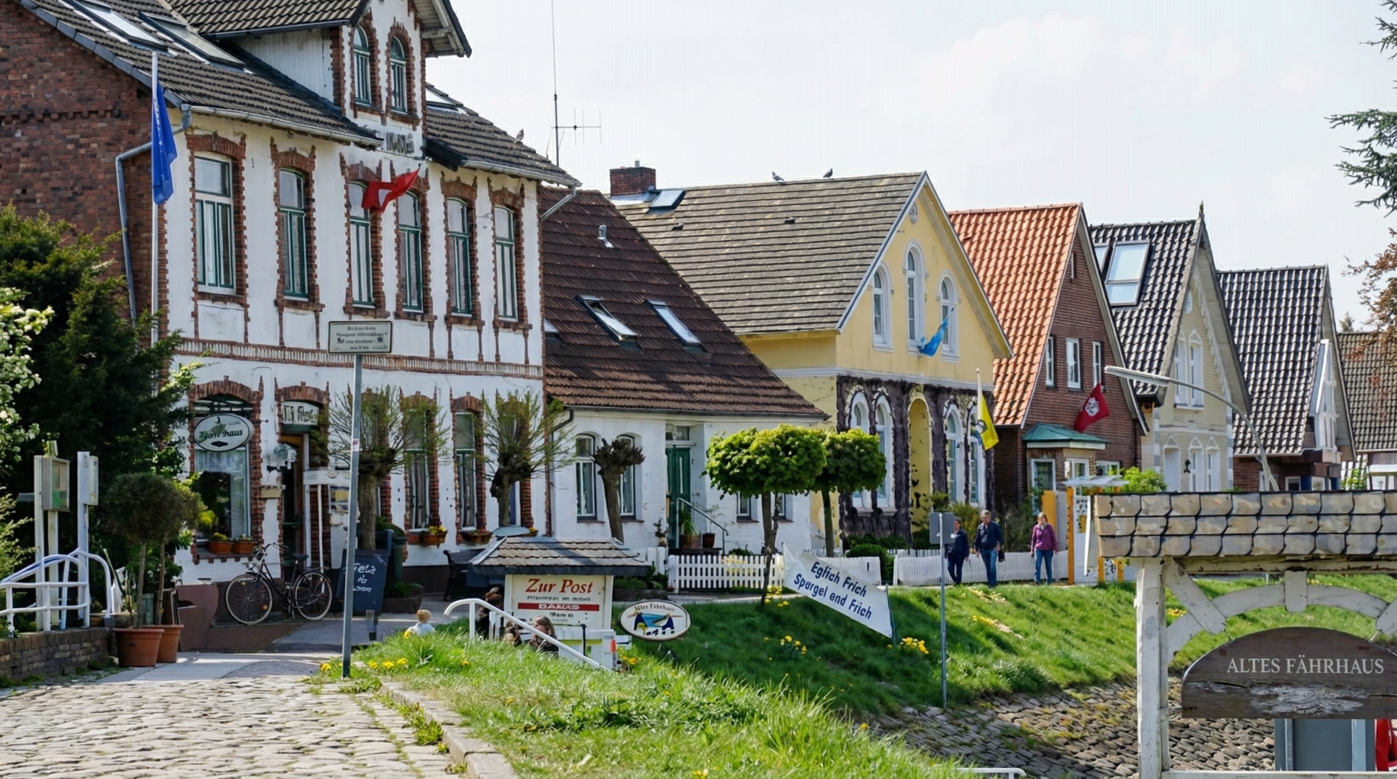 Reihe farbenfroher Häuser mit historischen Fassaden entlang einer gepflasterten Strasse am Wasser, darunter Gaststätten wie „Altes Fährhaus“ und „Zur Post“. Vor den Häusern stehen Schilder, Fahrräder und kleine Bäume, einige Menschen gehen spazieren in Cranz, Hamburg.
