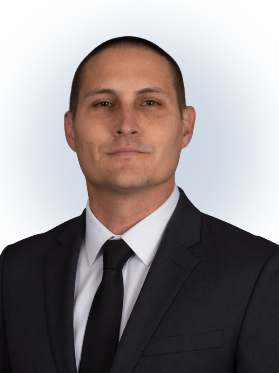 Man with short dark hair wearing a black suit, white shirt, and black tie against a light gradient background.
