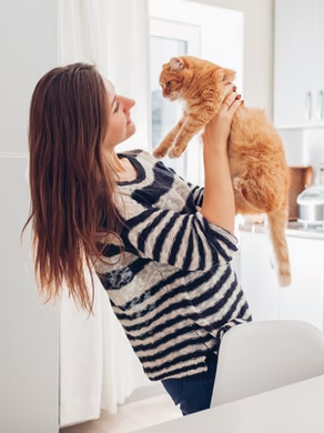 A woman holding an orange cat in her arms, smiling at it, in a modern kitchen setting.