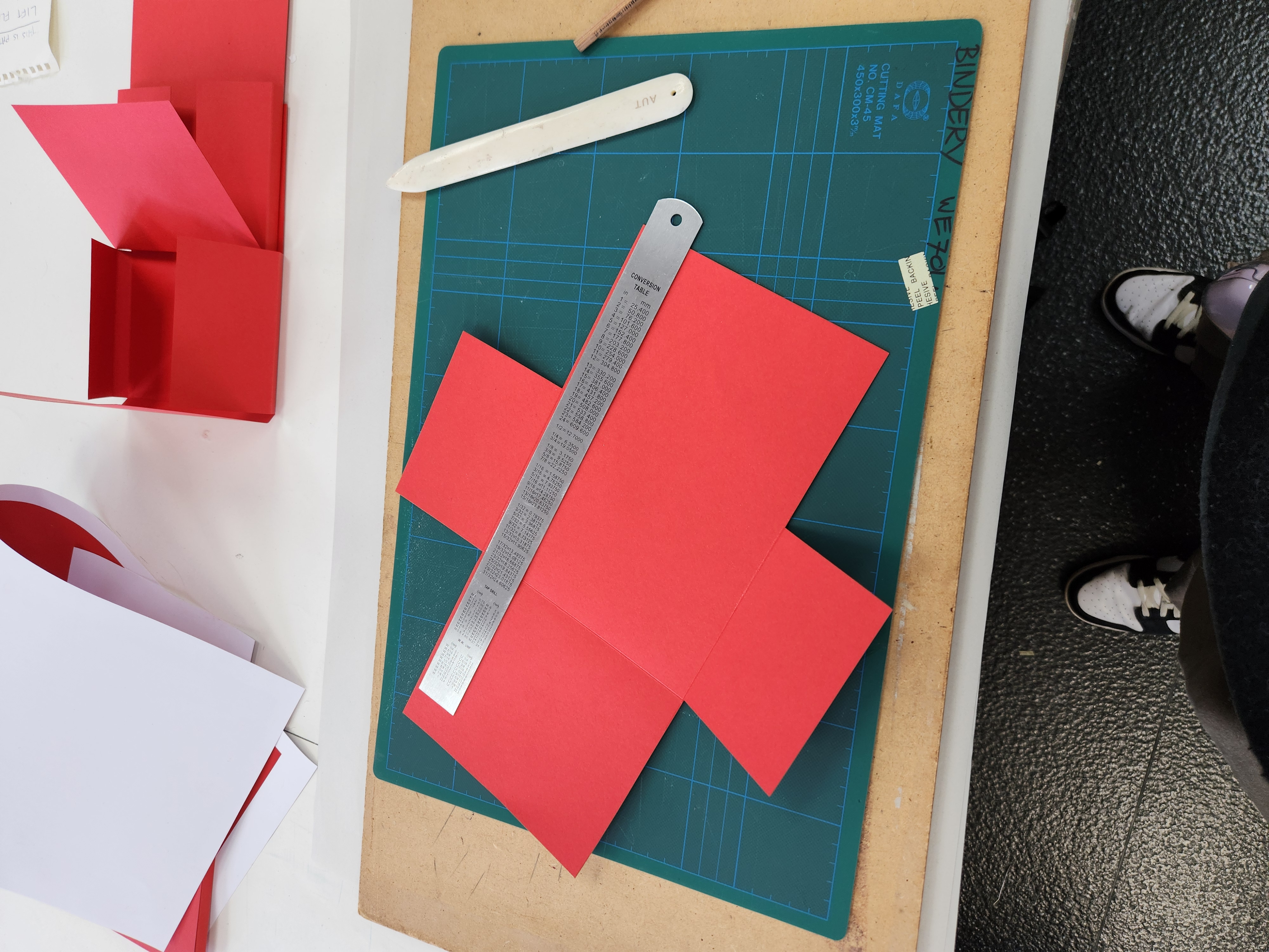 Red paper template on a green cutting mat with a metal ruler and bone folder, used for paper crafting.