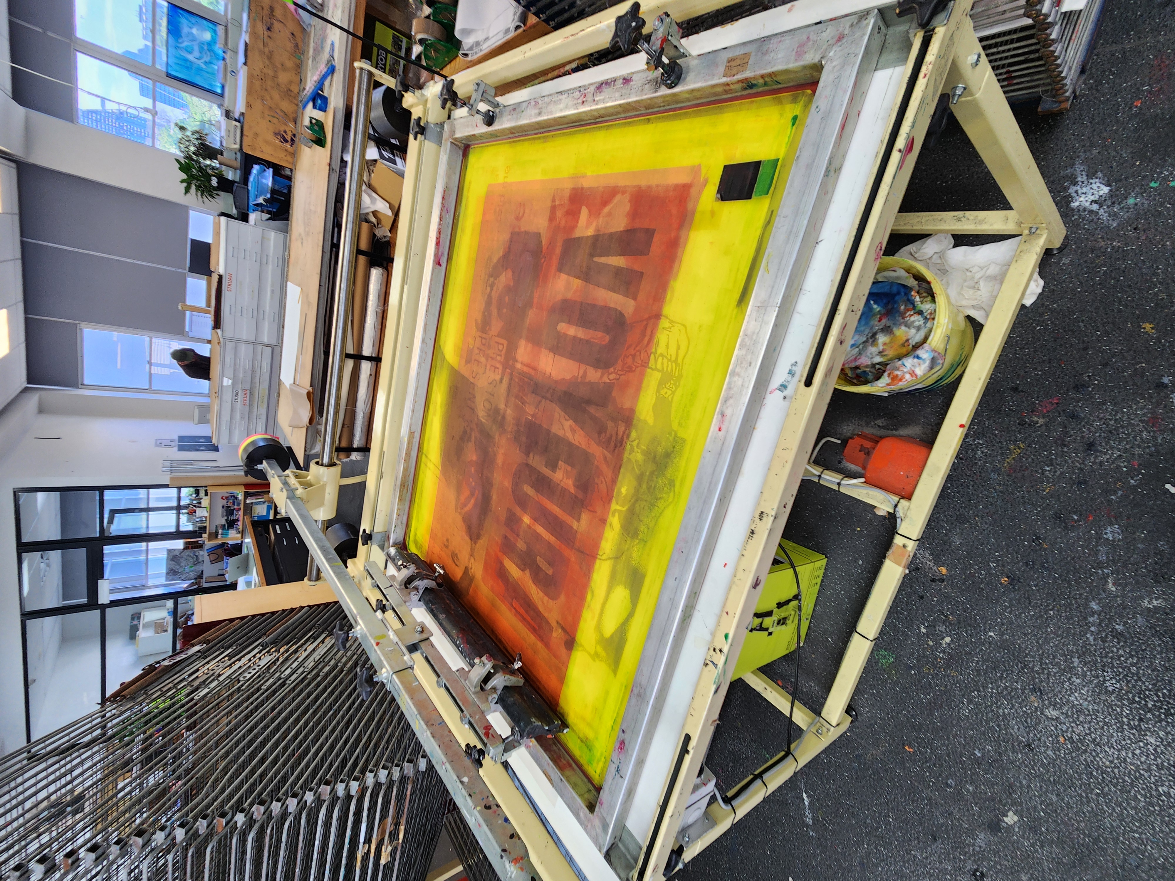 Screen printing table in a workshop with a yellow and orange screen showing reversed text and art supplies underneath.
