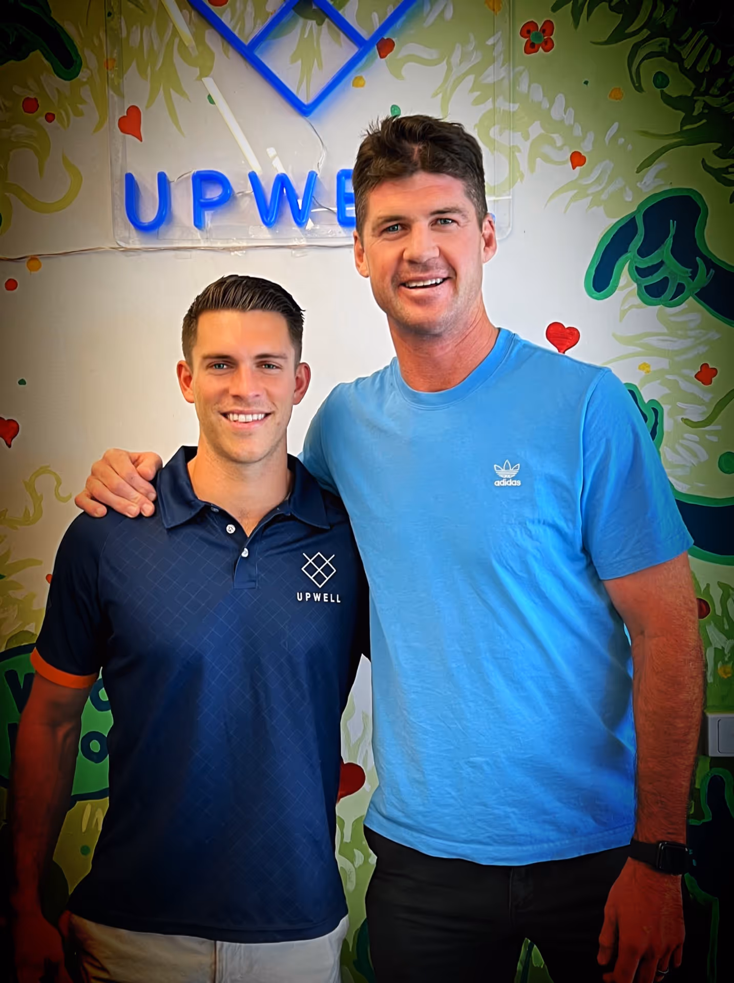Two smiling men standing side by side with one man's arm around the other's shoulder in front of a colorful wall with a neon sign that reads 'UPWELL'.
