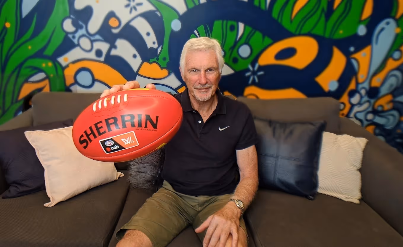 Older man with white hair sitting on a couch holding a red Sherrin Australian rules football.