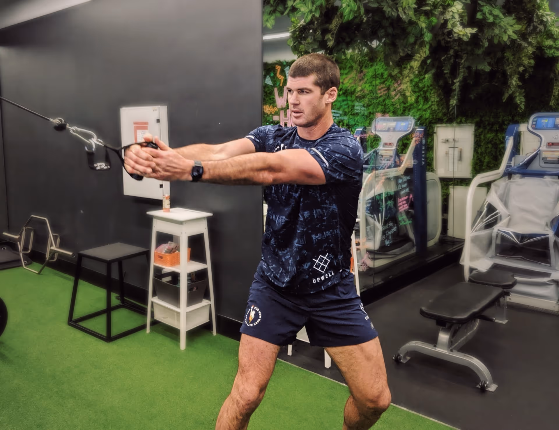 Man performing a cable workout exercise in a gym with green artificial turf and mirrored wall.
