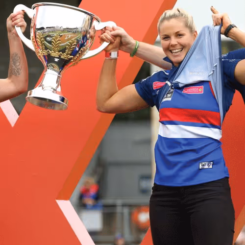 Smiling female athlete lifting a large silver trophy and holding a blue shirt, standing in front of a red geometric backdrop.