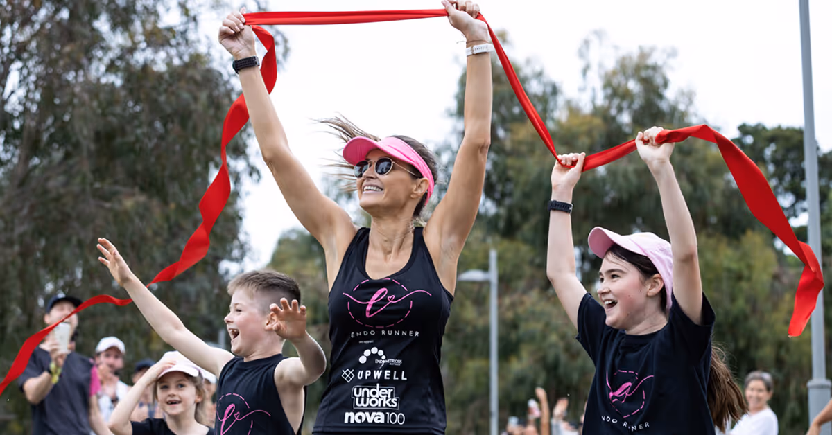 Smiling woman wearing sunglasses and pink visor holding a red finish-line ribbon with two children celebrating with raised arms during an outdoor event.