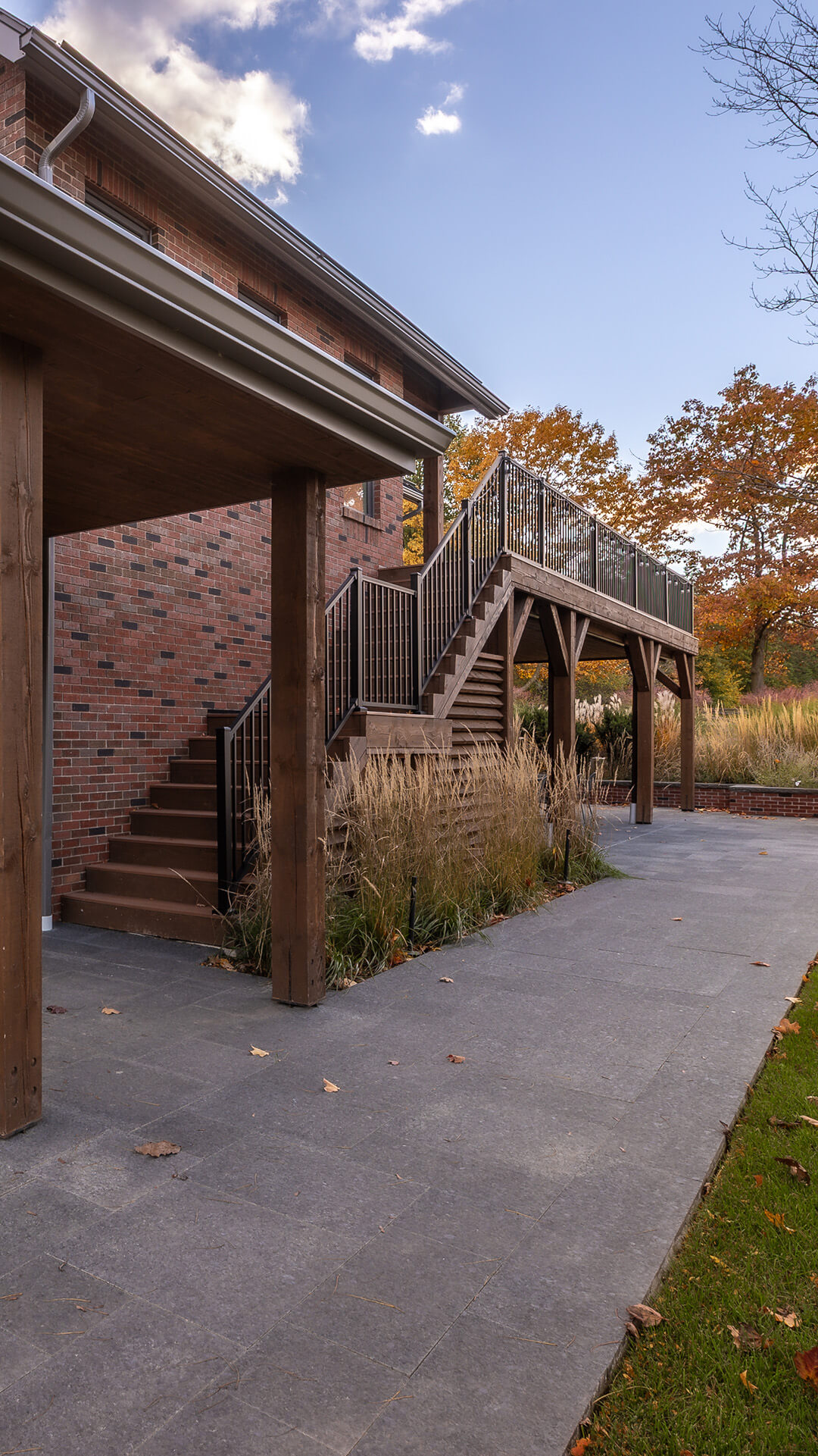Outdoor wooden staircase with black metal railing leading to a raised deck attached to a brick house, surrounded by autumn foliage and ornamental grasses.