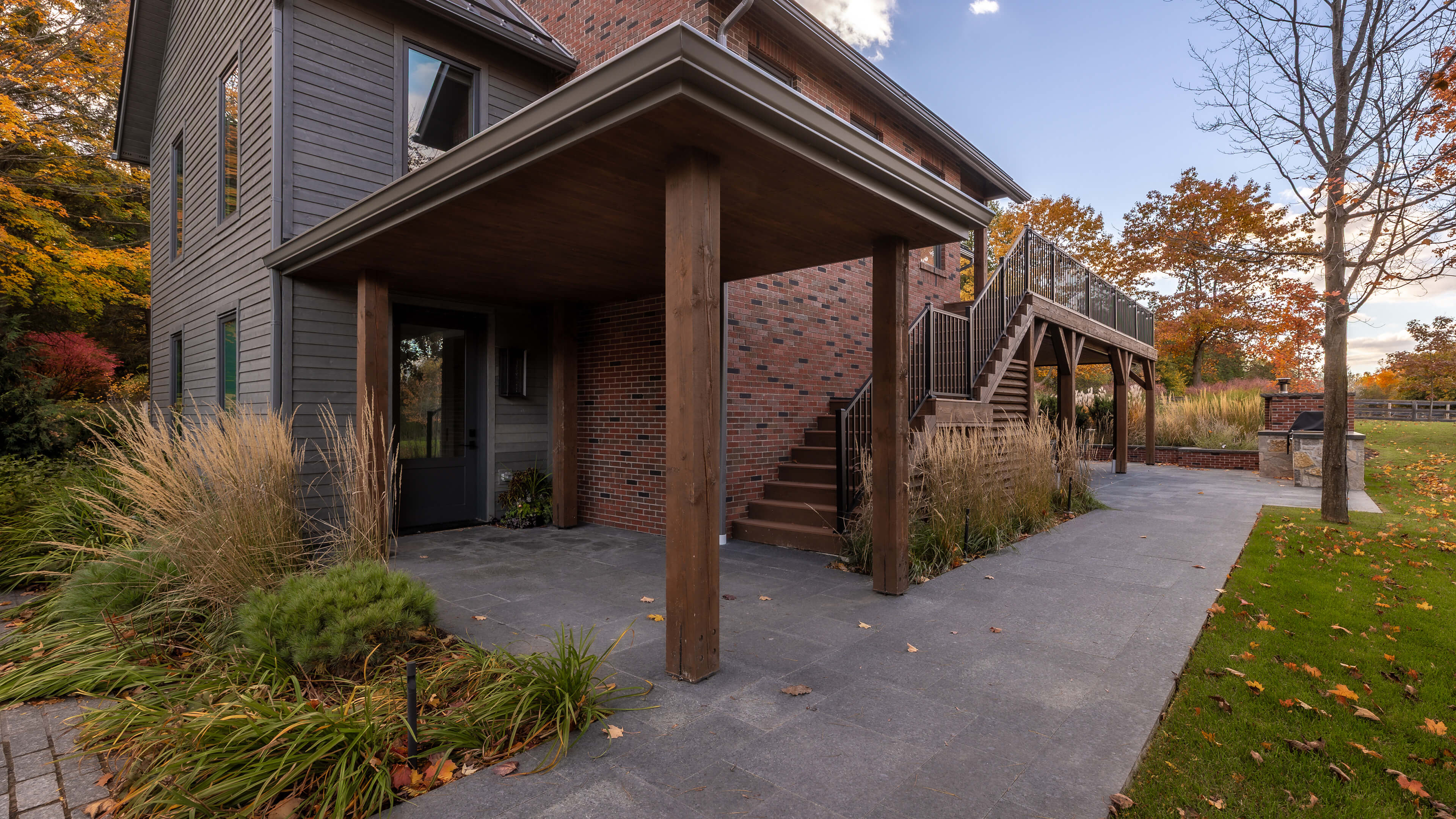 Backyard patio with brick and wood house exterior, stairs leading to elevated deck, autumn trees, and outdoor grill area.