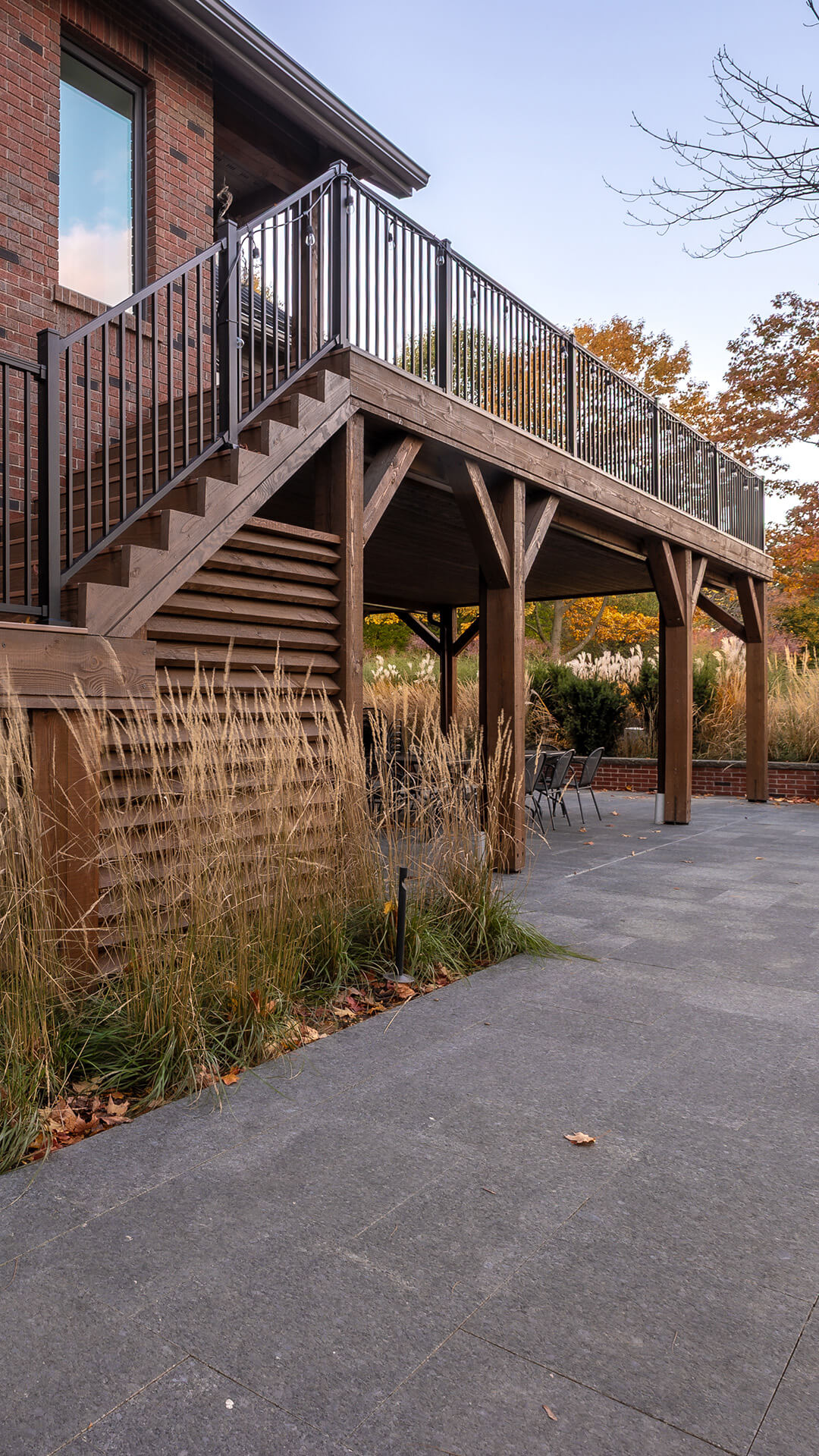 Wooden raised deck with black metal railing and stairs next to a brick house, overlooking a gray stone patio and autumn foliage.