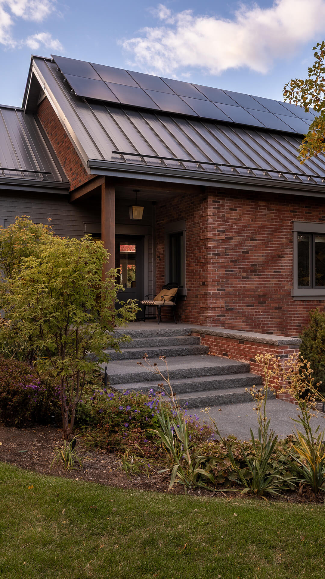 Brick house with a metal roof featuring solar panels, steps leading to a porch with a bench and plants in the garden.