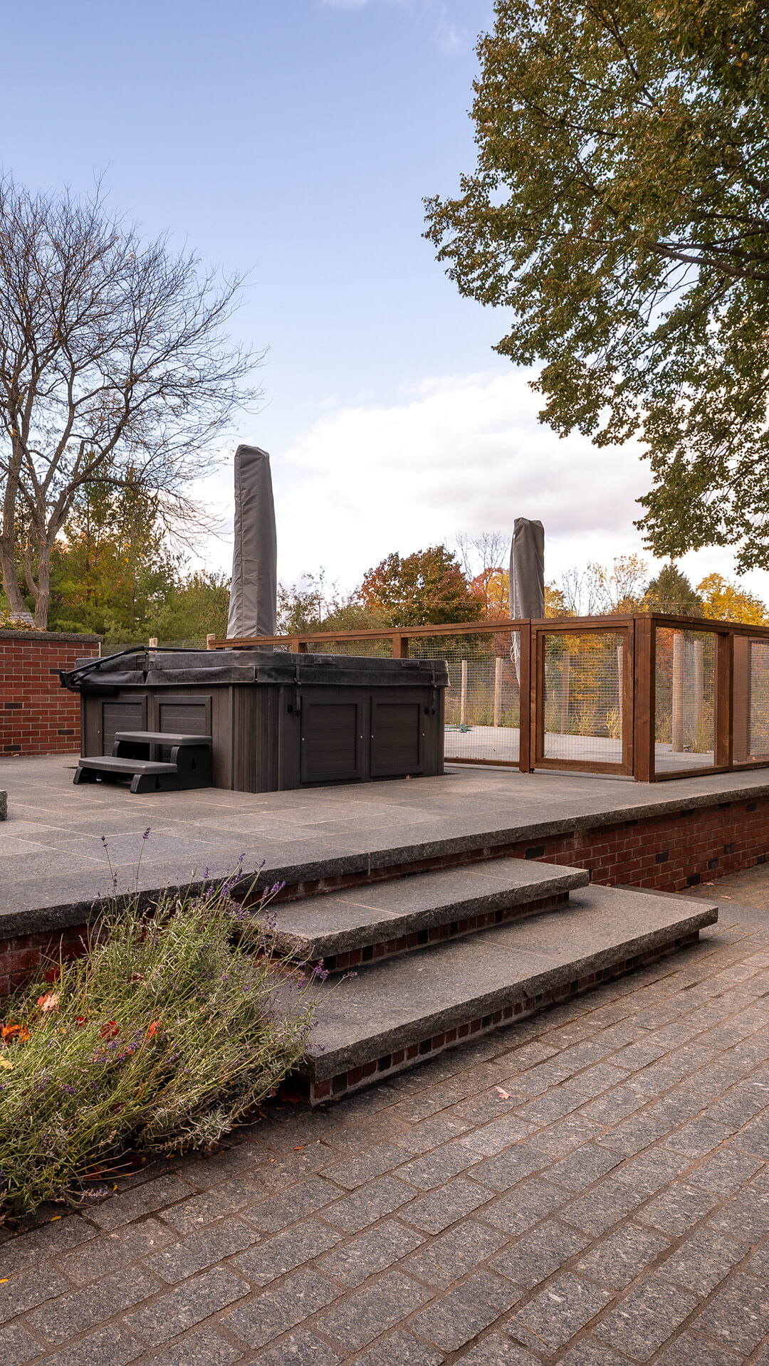 Outdoor hot tub with black cover and small black steps on raised stone patio surrounded by wooden fence, with trees and cloudy sky in background.
