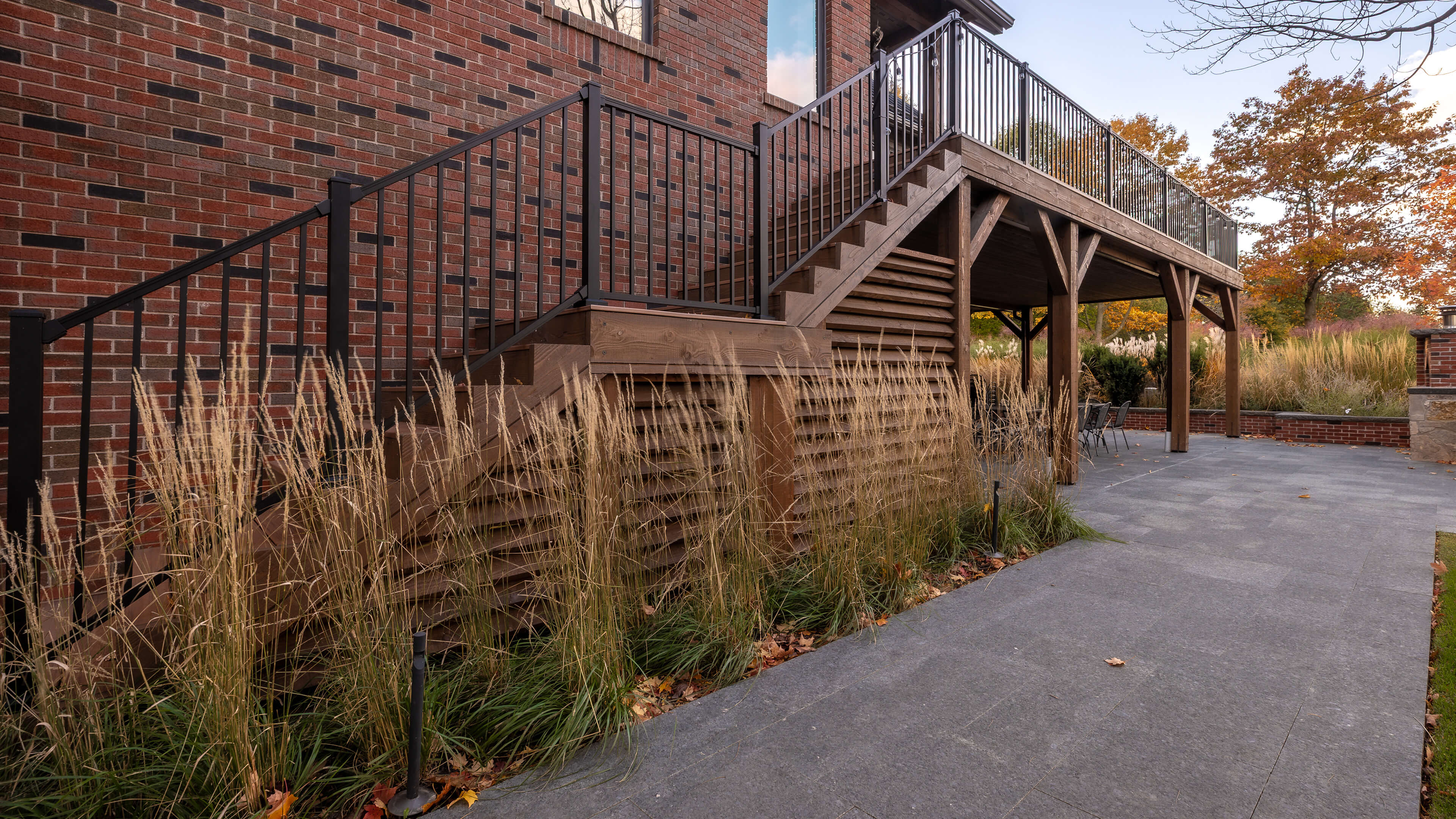 Wooden outdoor staircase with black metal railings attached to a brick house, surrounded by ornamental grasses and leading to an elevated deck with fall foliage in the background.
