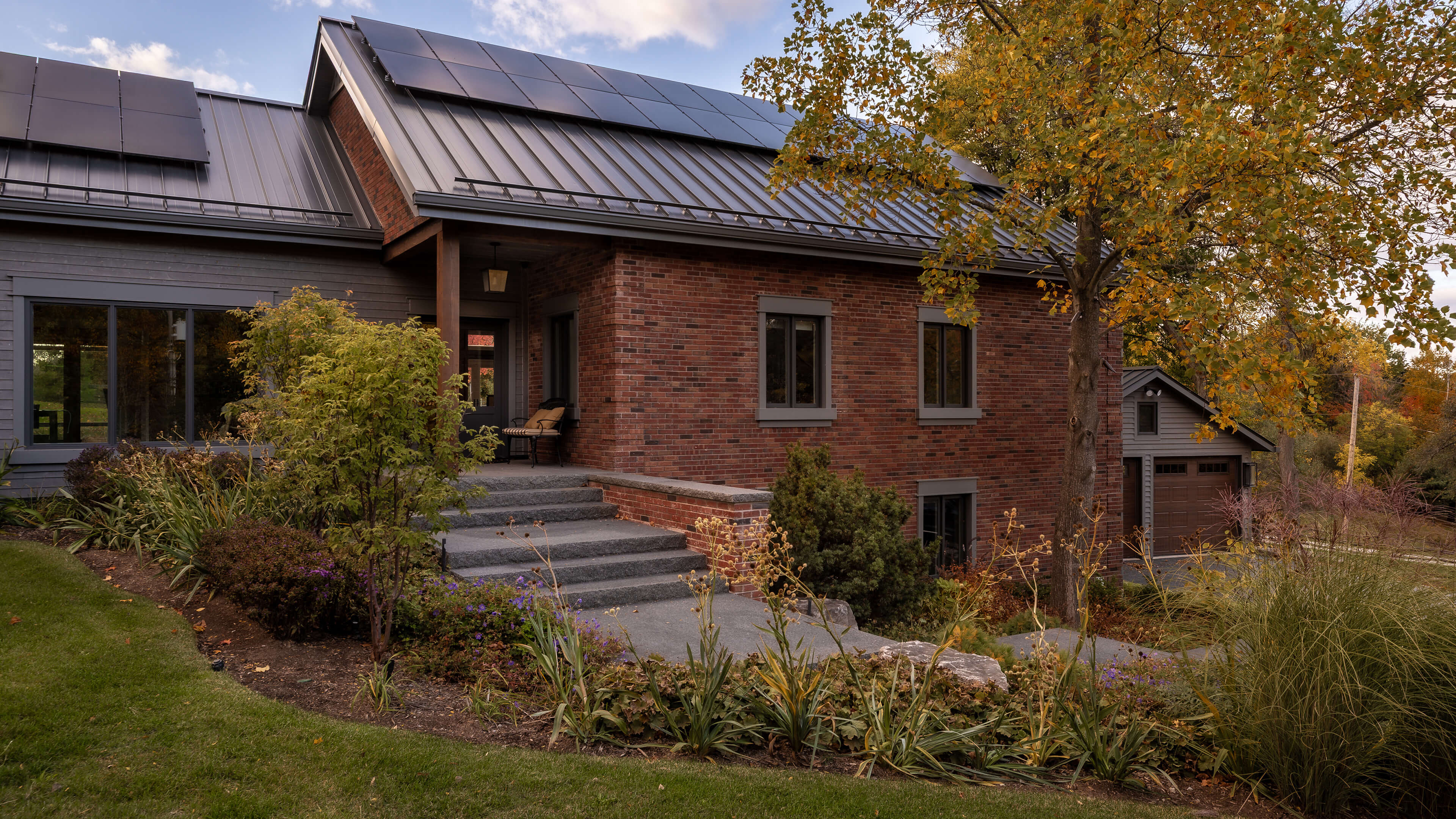 Modern brick and gray wood house with solar panels on roof, surrounded by autumnal landscaping and a large tree.