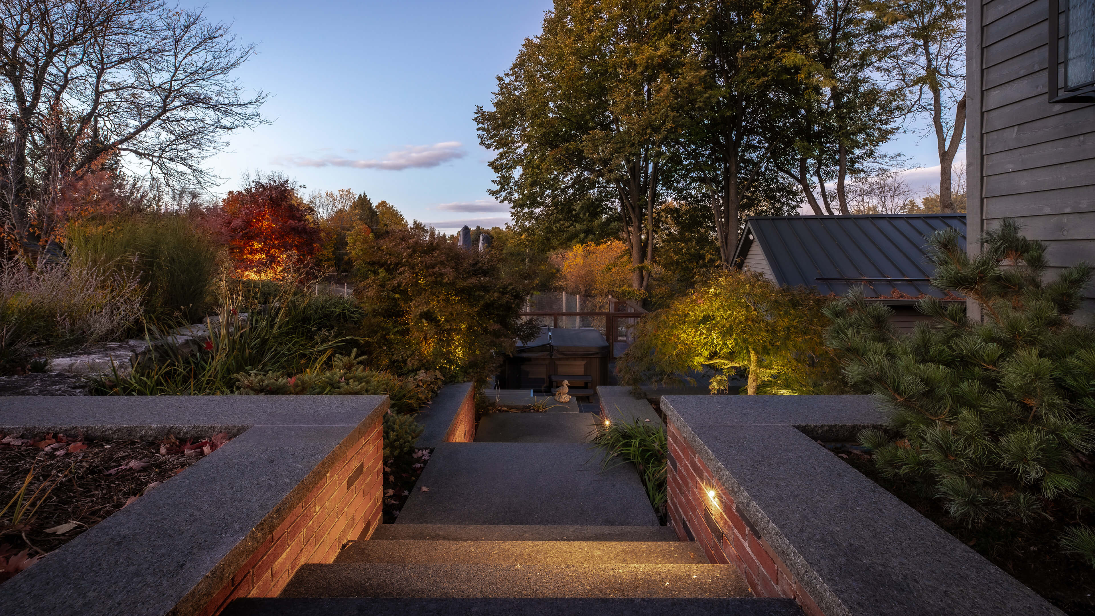 Stone steps with brick sidewalls leading down to a garden area with illuminated bushes and large trees under a clear evening sky.
