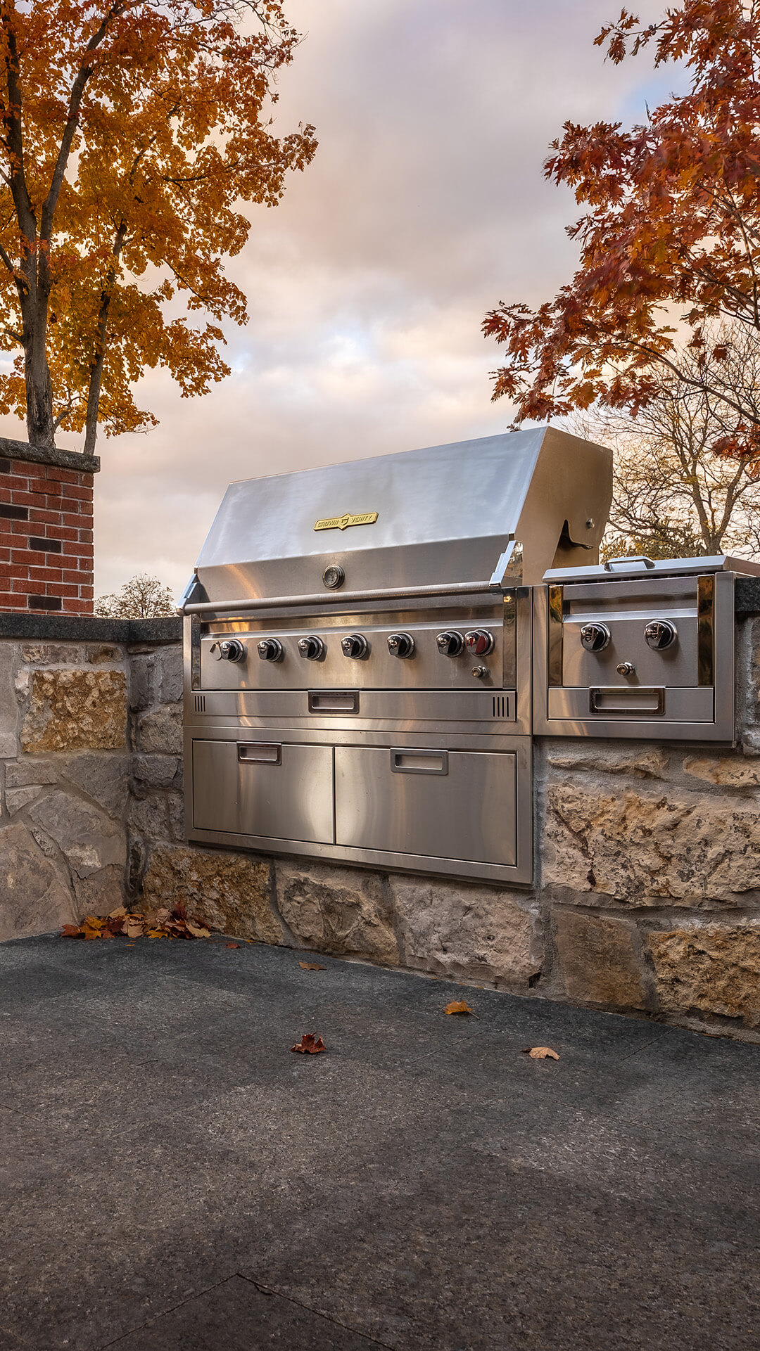 Built-in stainless steel outdoor grill set into stone wall with autumn trees and cloudy sky background.