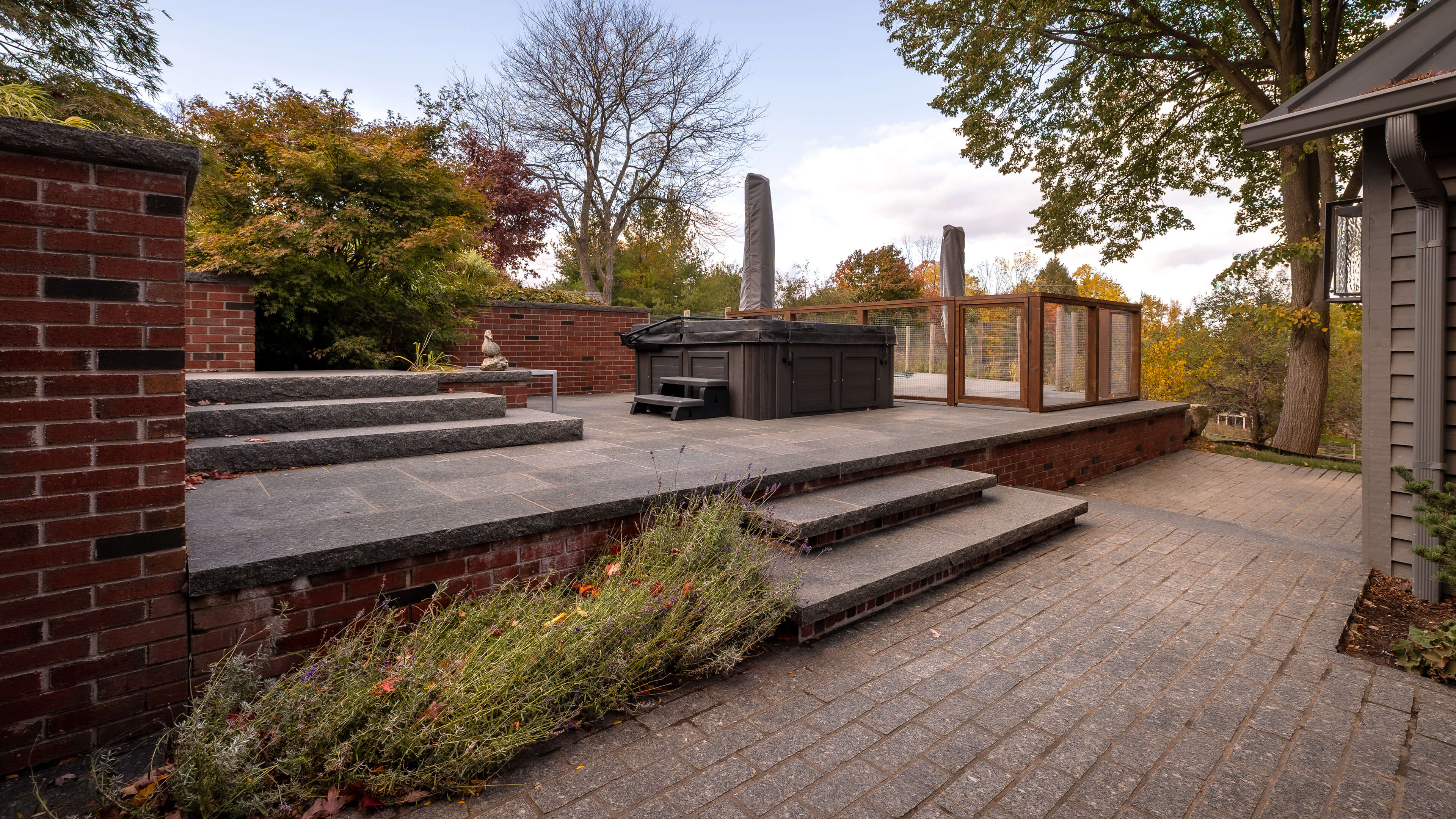 Raised stone patio with a covered hot tub, surrounded by brick walls and autumn trees.