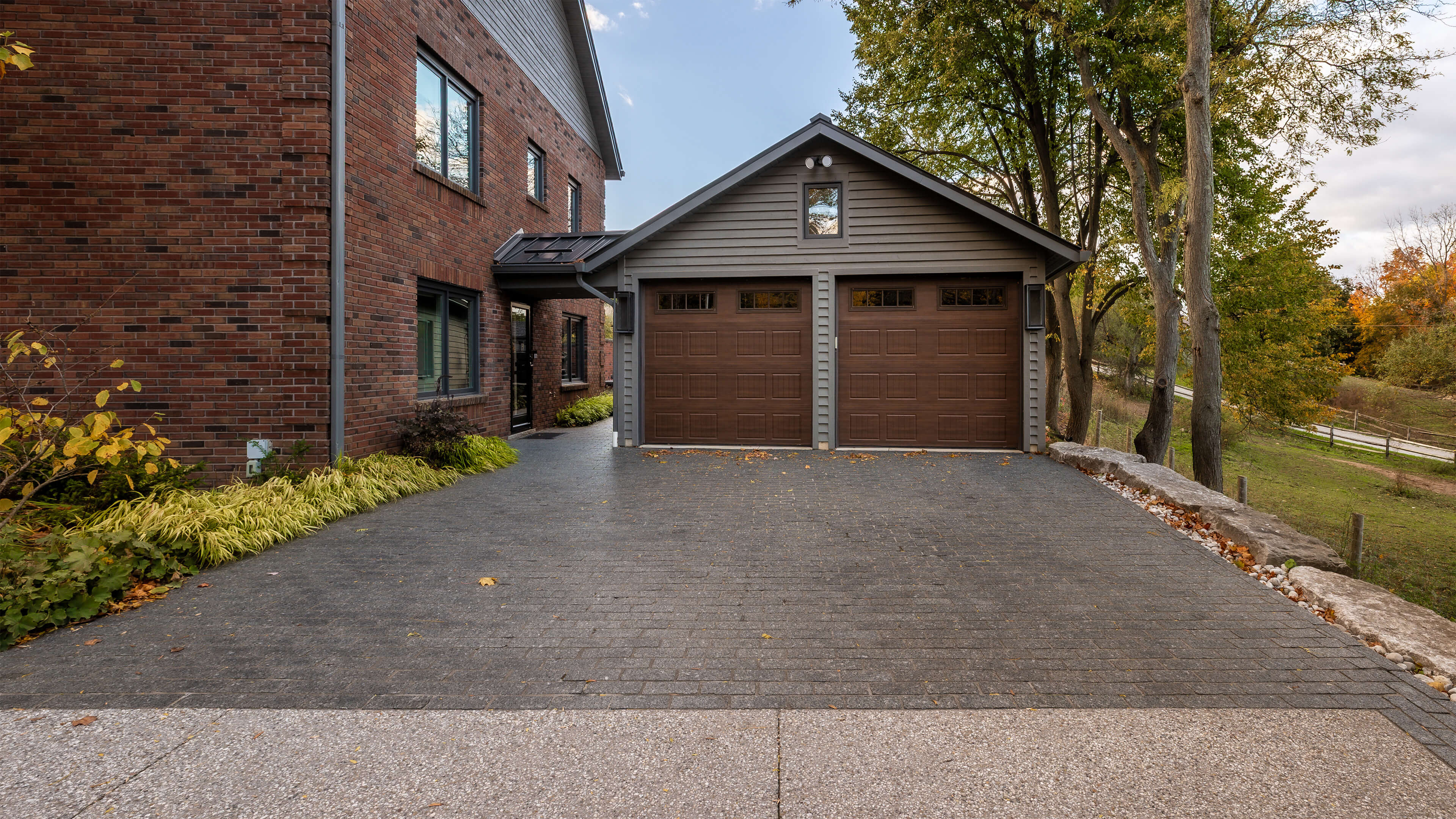 Driveway leading to a detached garage with two wooden doors, adjacent to a brick house and surrounded by trees in autumn.