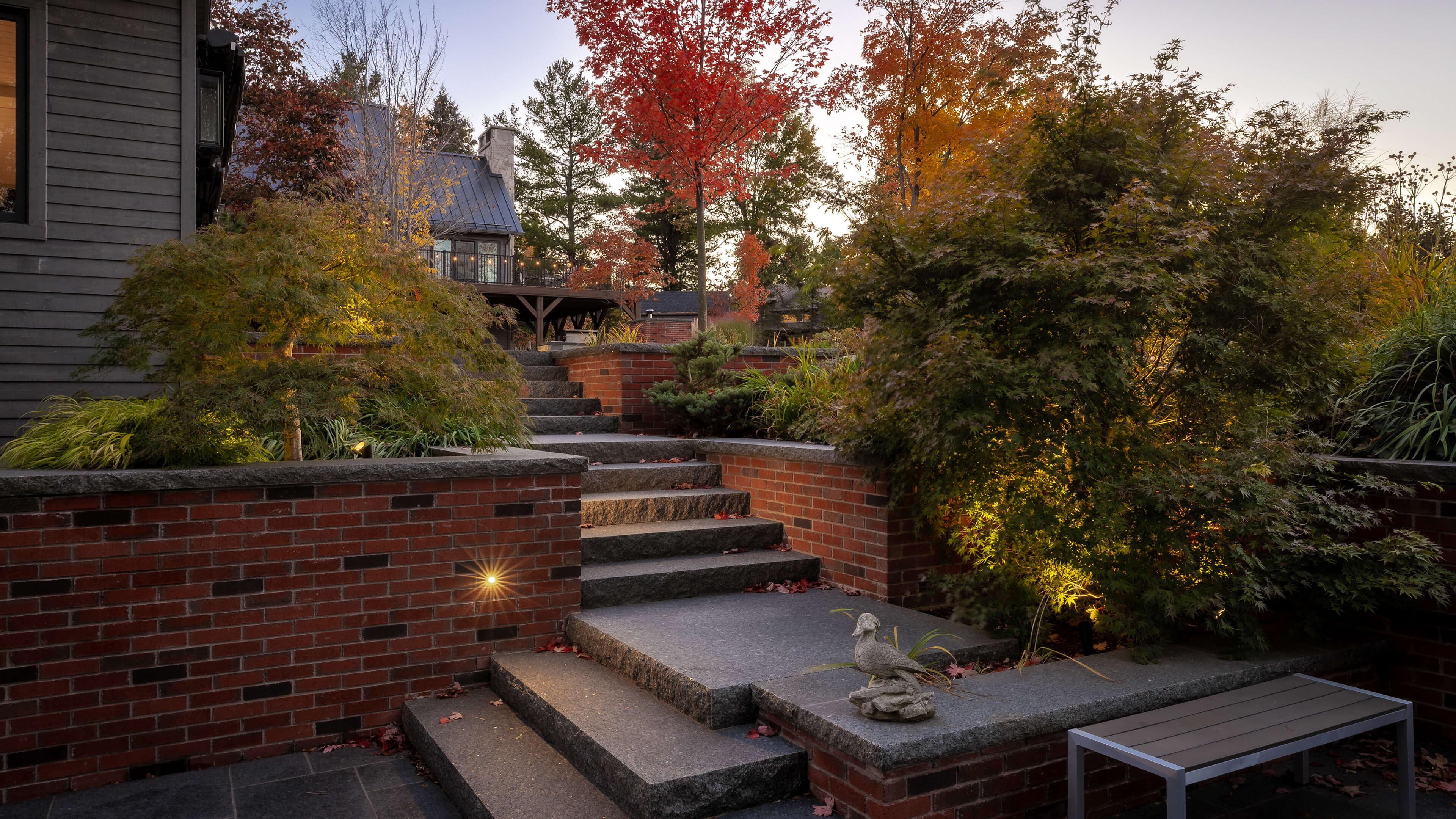 Outdoor stone steps with brick walls, illuminated small trees with autumn leaves, a stone duck sculpture, and a wooden bench at dusk.