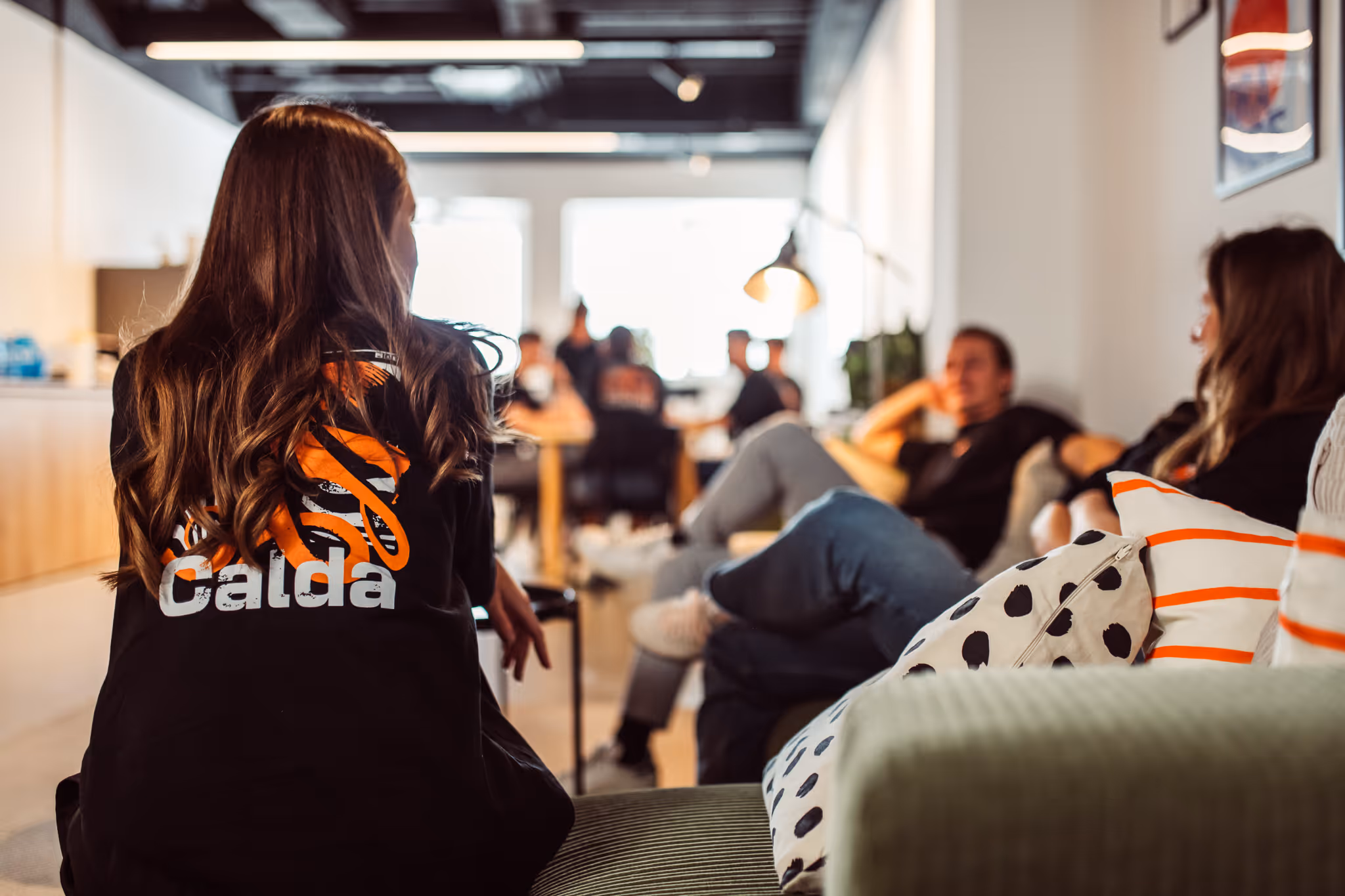 People relaxing and talking in a casual office lounge with one person wearing a black jacket with 'Calda' logo.