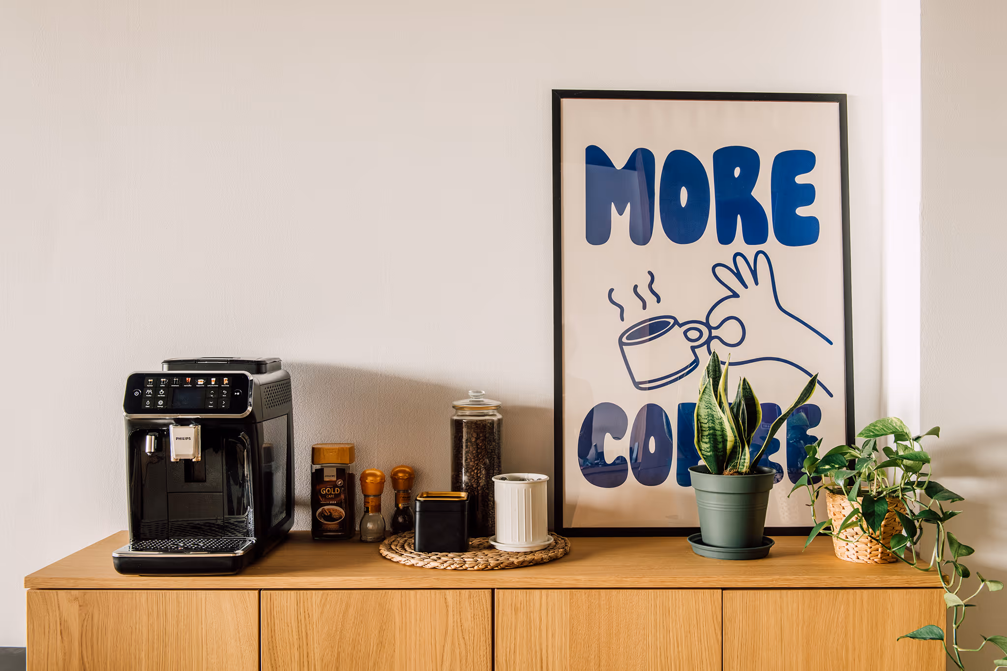 Coffee corner with a black coffee machine, jars of coffee beans and spices, two potted plants, and a framed print reading 'MORE COFFEE' on a wooden sideboard.