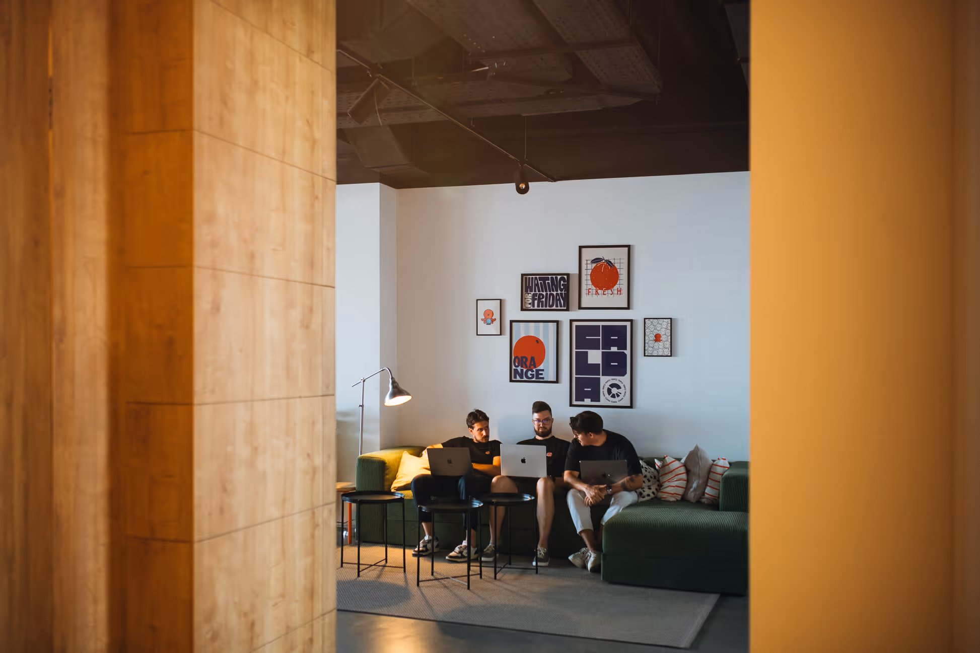 Three men sitting on a green couch with laptops, working together in a modern office space with framed artwork on the wall.