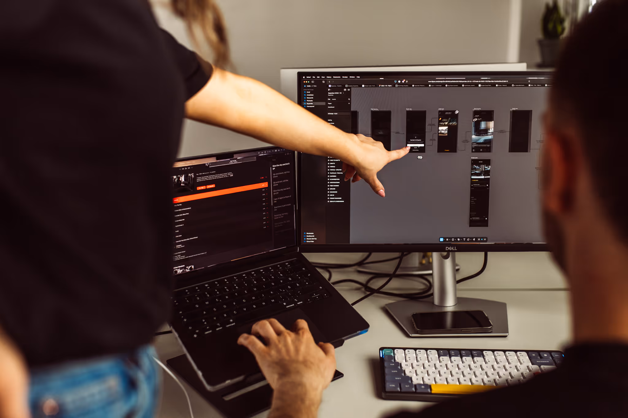 Person pointing at a computer monitor displaying a design interface while another person uses a laptop keyboard.