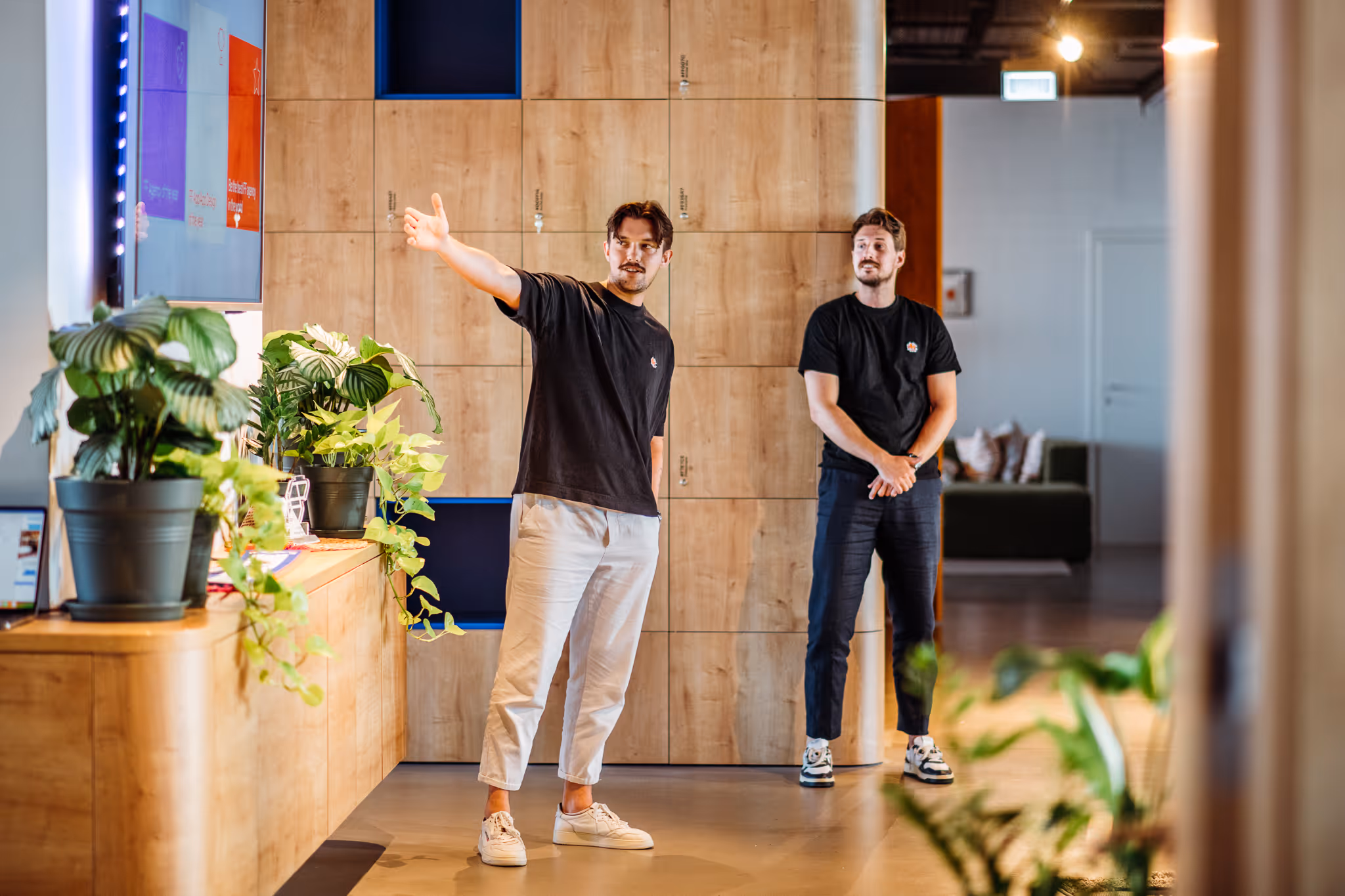 Two men in black t-shirts standing indoors by wooden lockers, one gesturing toward a screen on the wall.