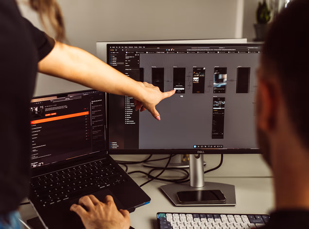 Person pointing at a desktop monitor displaying app design wireframes while another uses a laptop.