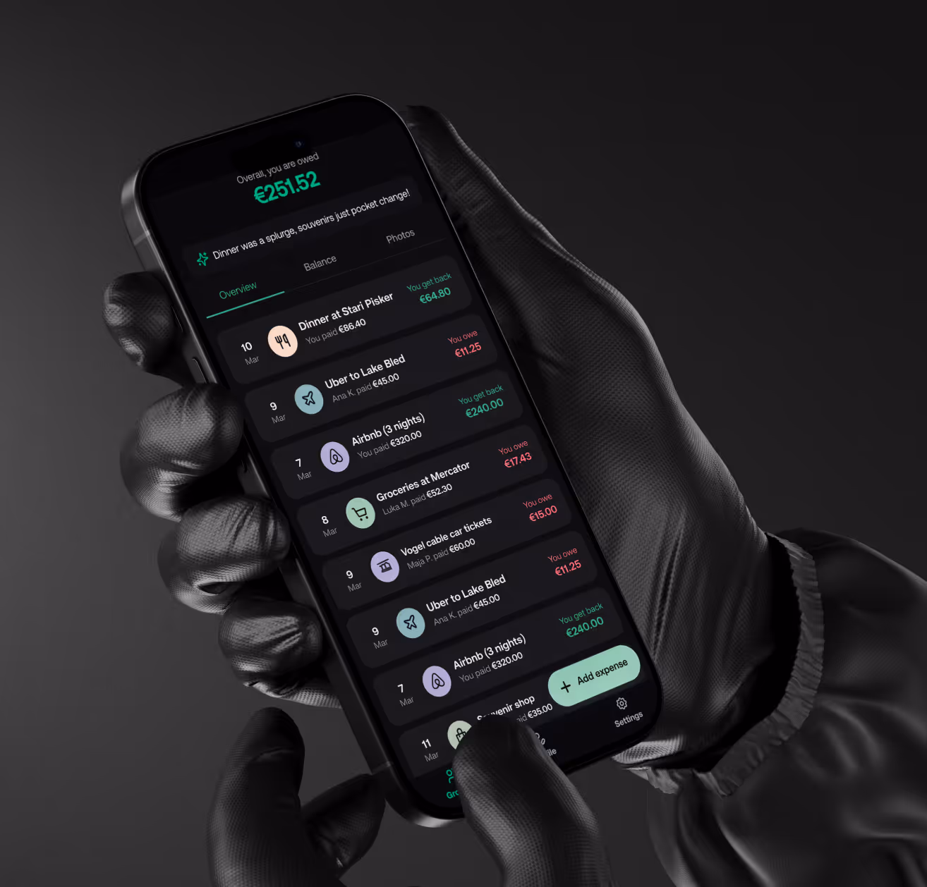Person wearing black gloves holding a phone showing a dark-mode expense tracking app with balances.