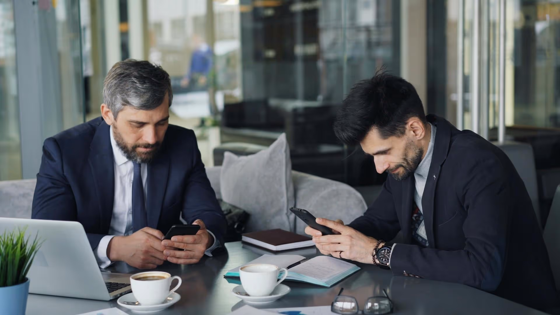 Two businessmen in suits sitting at a modern office table, focused on their smartphones. The table features notebooks, a laptop, and coffee cups, with a professional environment visible in the background.