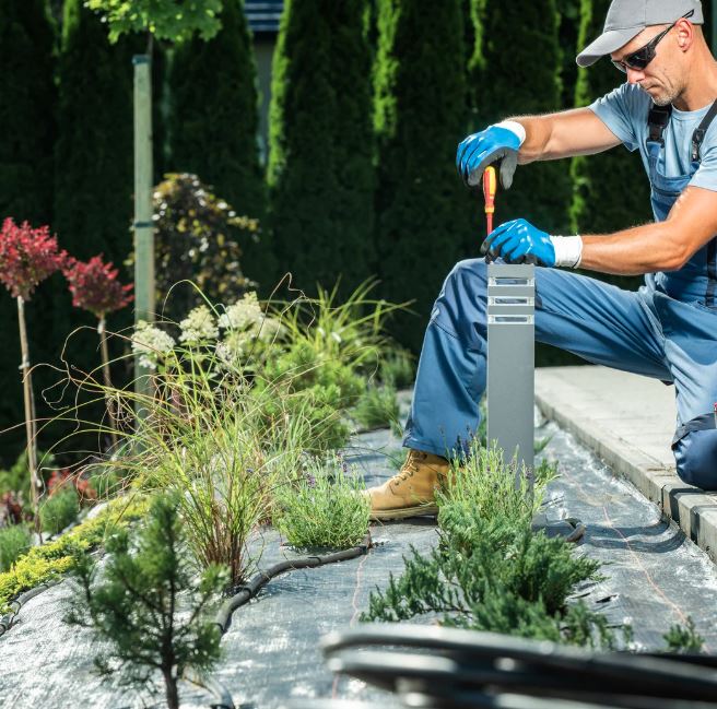 Worker performing low-voltage outdoor lighting upgrade in landscaped garden for energy-efficient illumination.