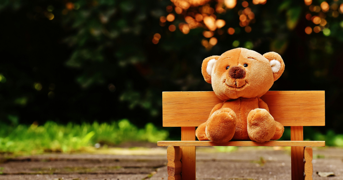 Ours en peluche vintage posé sur un banc en plein air sous un ciel ensoleillé — objet de collection apprécié des passionnés de peluches et jouets anciens.