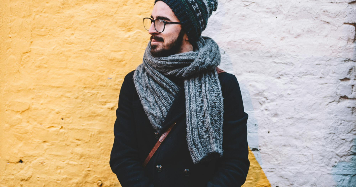homme qui a plus de la trentaine, avec une barbe brune. Il porte des lunettes, un bonnet et une écharpe. Adossé à un mur colorés, il regarde de côté.