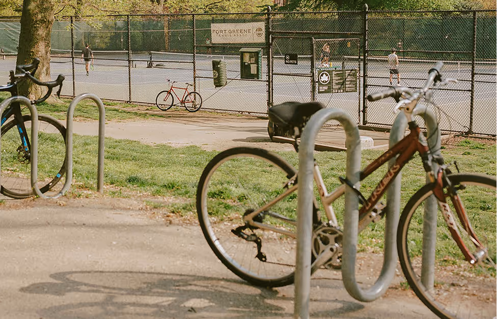 Bicycles locked to racks near a fenced tennis court with people playing in a park.