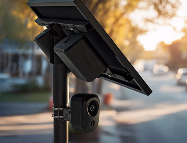 Close-up of a solar-powered security camera mounted on a pole with a street in the background.