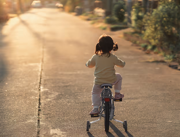 Young child riding a bicycle with training wheels on a sunlit road in the late afternoon.