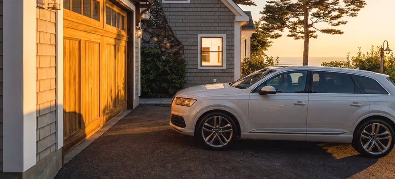 Silver SUV parked on driveway in front of a house with wooden garage door during sunset.