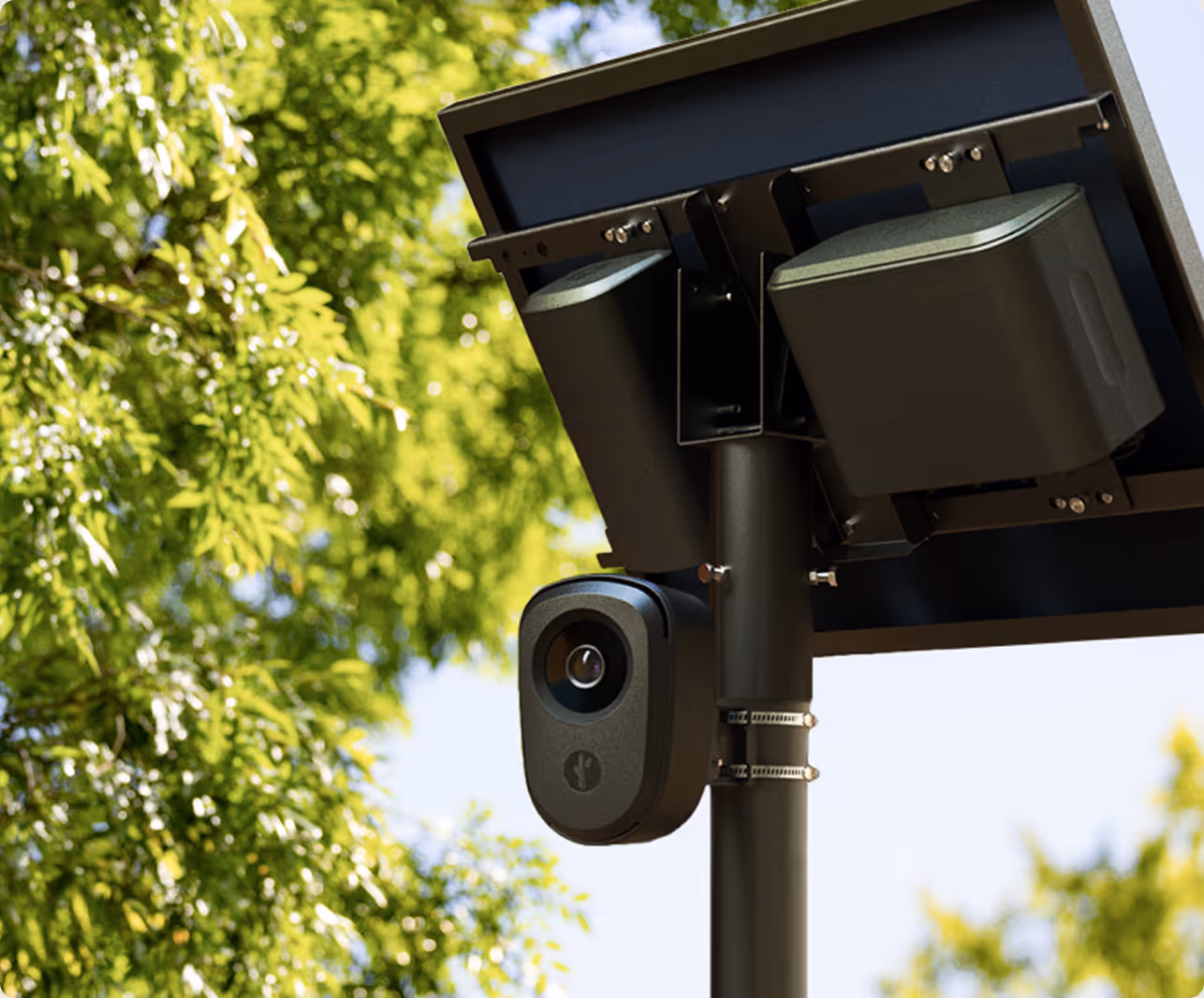 Black security camera mounted on a pole with a solar panel and green leafy trees in the background.